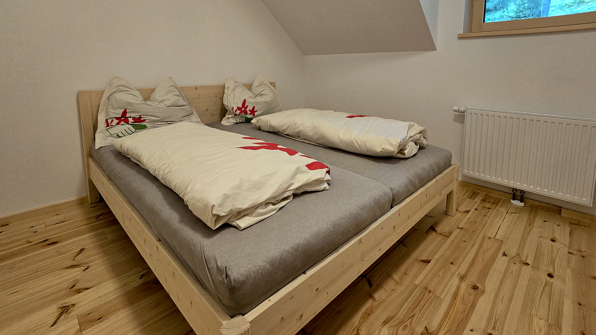 Ein ordentlich gemachtes Doppelbett in der Bleckwandh&uuml;tte mit einem hellen Holzrahmen, grauen Laken und cremefarbener Bettw&auml;sche in einem schlichten Zimmer.