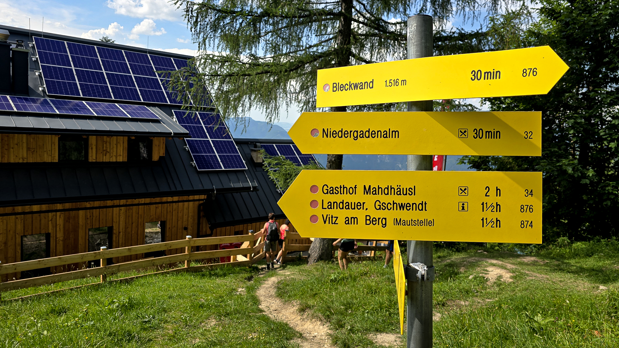 Gelbe Wanderwegweiser mit Zeitangaben in der N&auml;he der Bleckwandh&uuml;tte, einem Holzgeb&auml;ude mit Solaranlage in den Bergen.