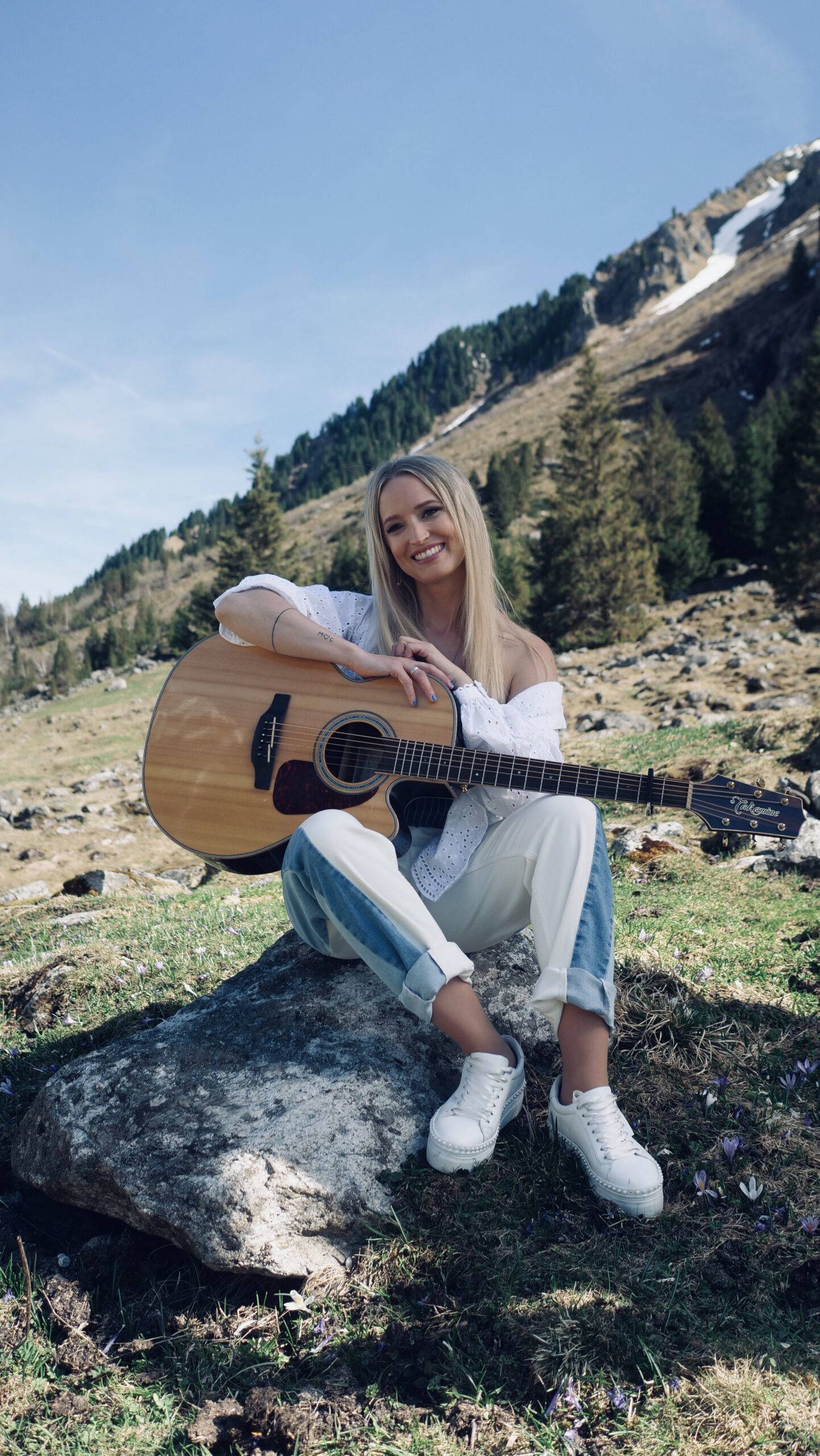 Eine lächelnde Frau sitzt draußen auf einem Felsen und hält eine Akustikgitarre in der Hand, im Hintergrund sind Berge und Bäume zu sehen.