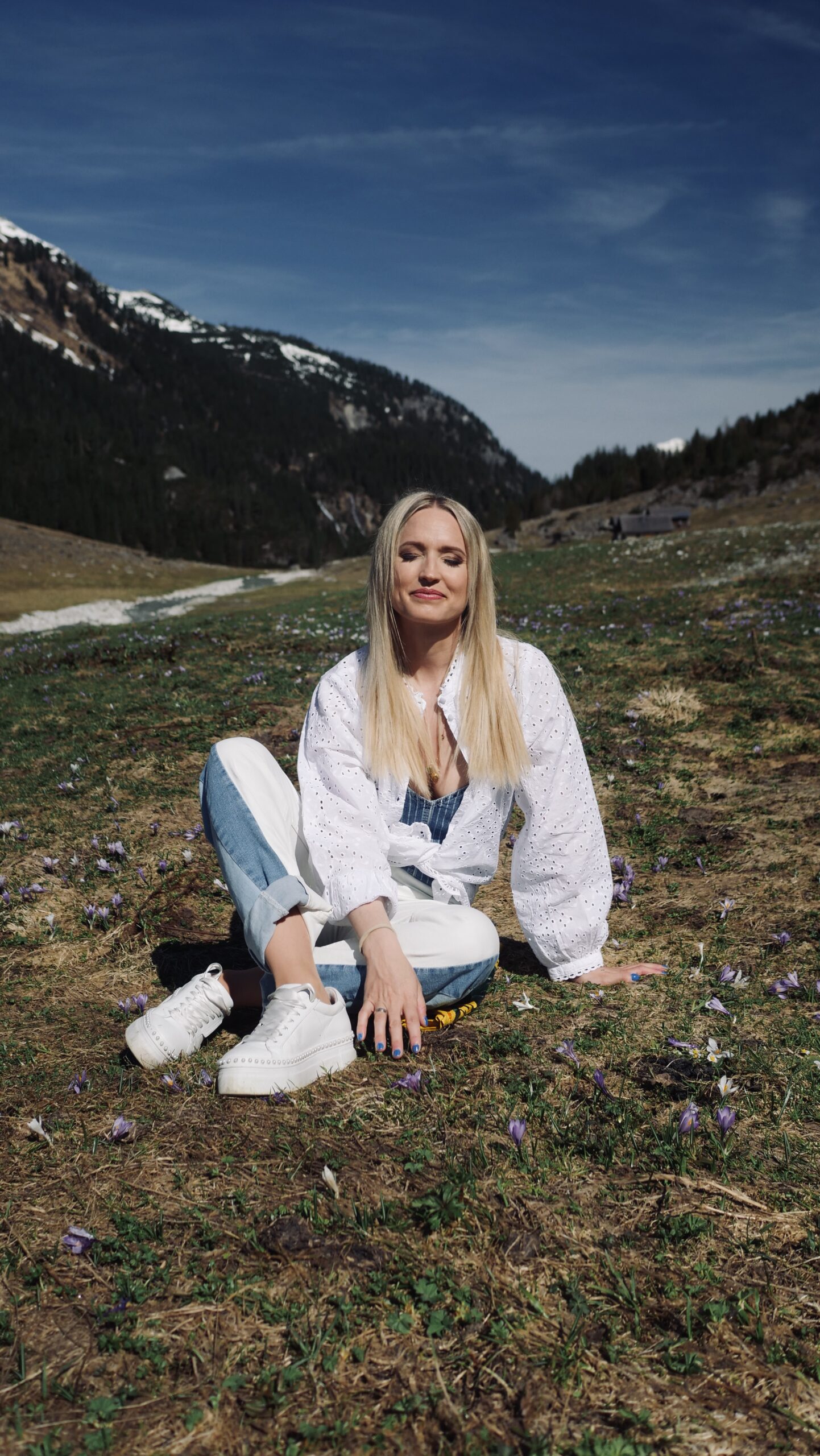 Eine Frau mit langen blonden Haaren sitzt auf dem Gras einer Bergwiese, umgeben von violetten Blumen und schneebedeckten Gipfeln.