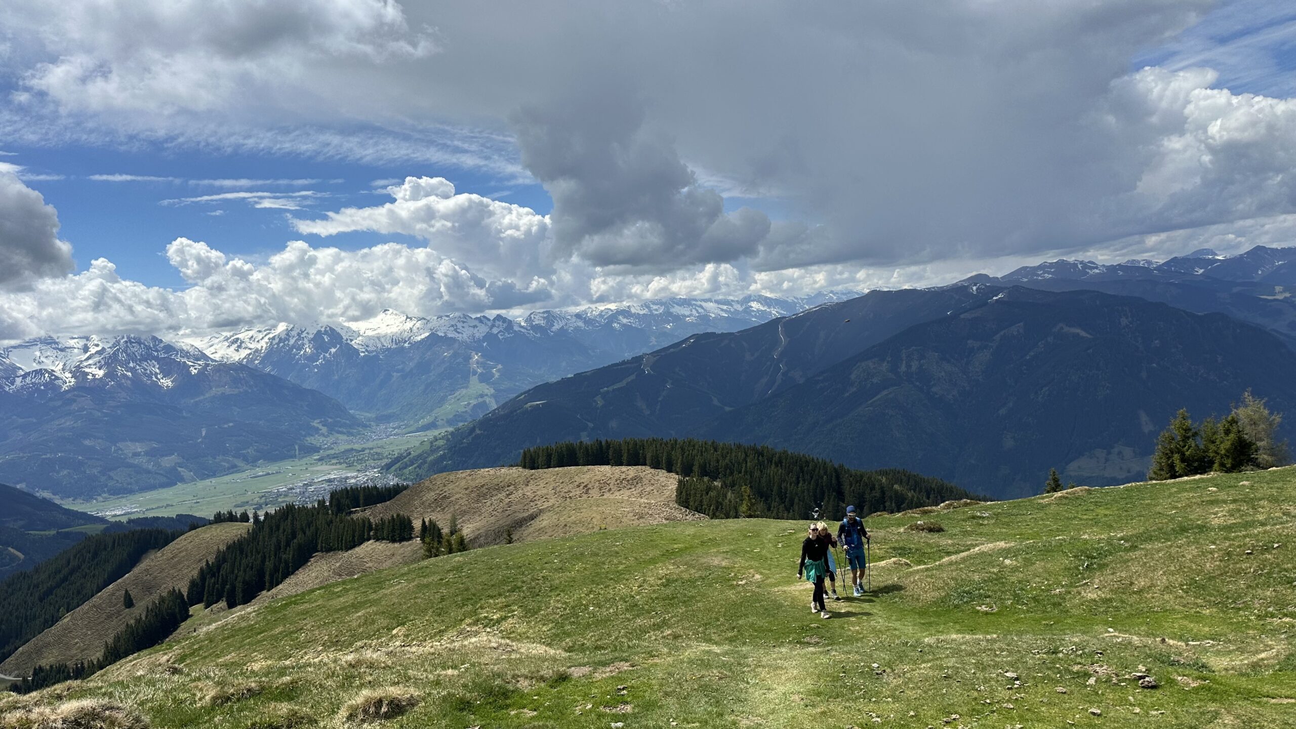 Zwei Wanderer gehen auf einem grasbewachsenen Weg auf der Schwalbenwand, im Hintergrund sind schneebedeckte Gipfel und dramatische Wolken zu sehen.
