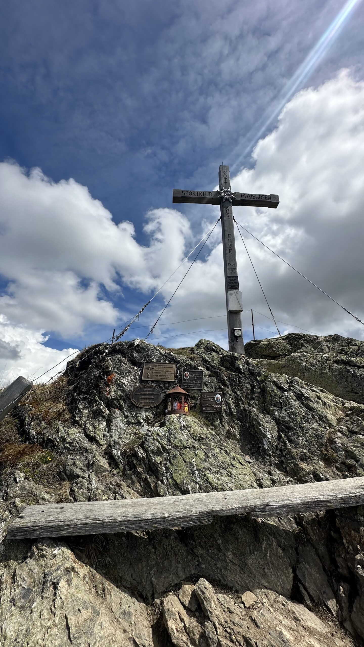 Ein großes Holzkreuz thront auf dem felsigen Gipfel der Schwalbenwand, darunter befinden sich Gedenktafeln unter einem bewölkten Himmel.