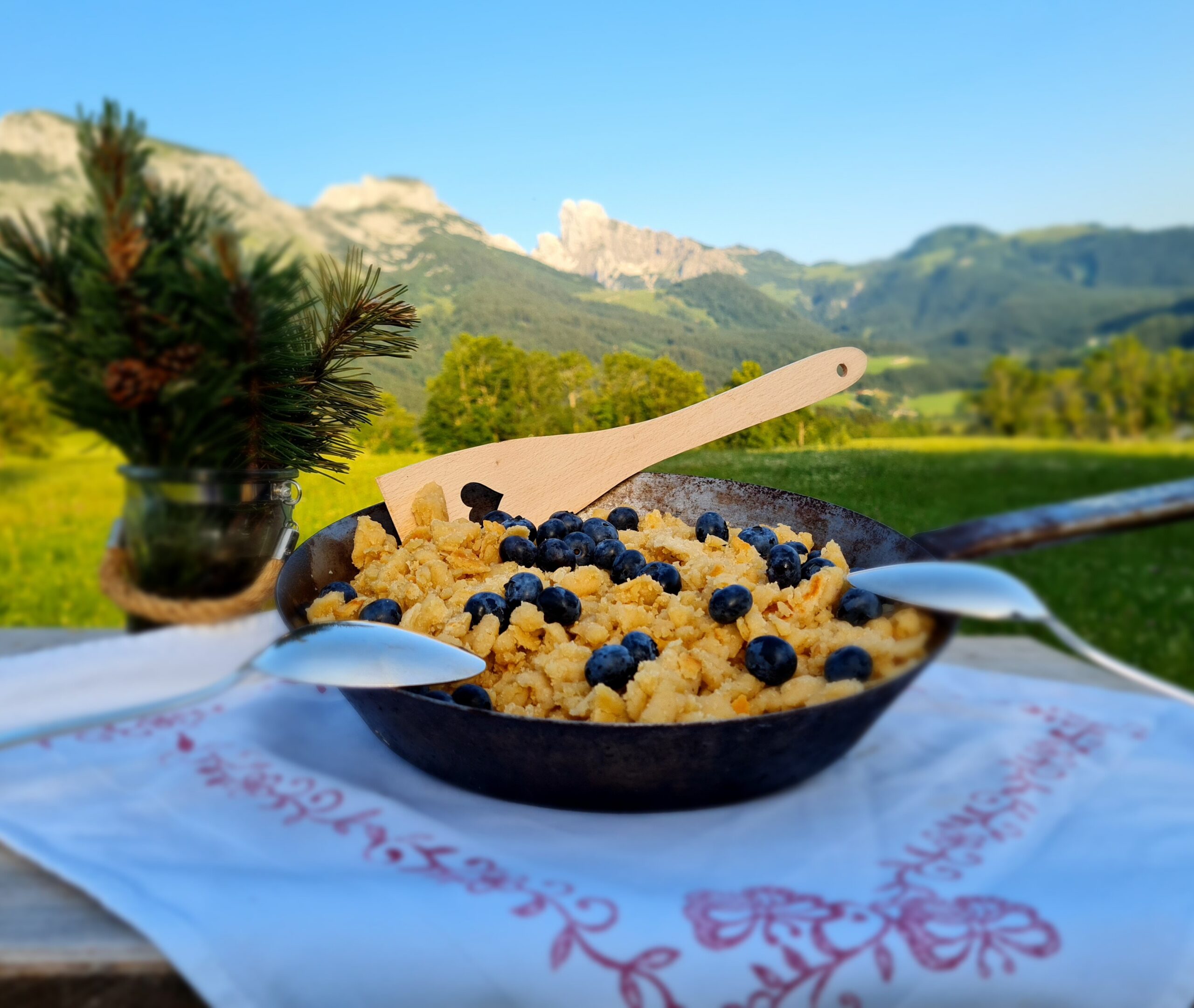 Auf einem Tisch im Freien steht eine Pfanne mit Rührei und Heidelbeeren, im Hintergrund sind Berge zu sehen.