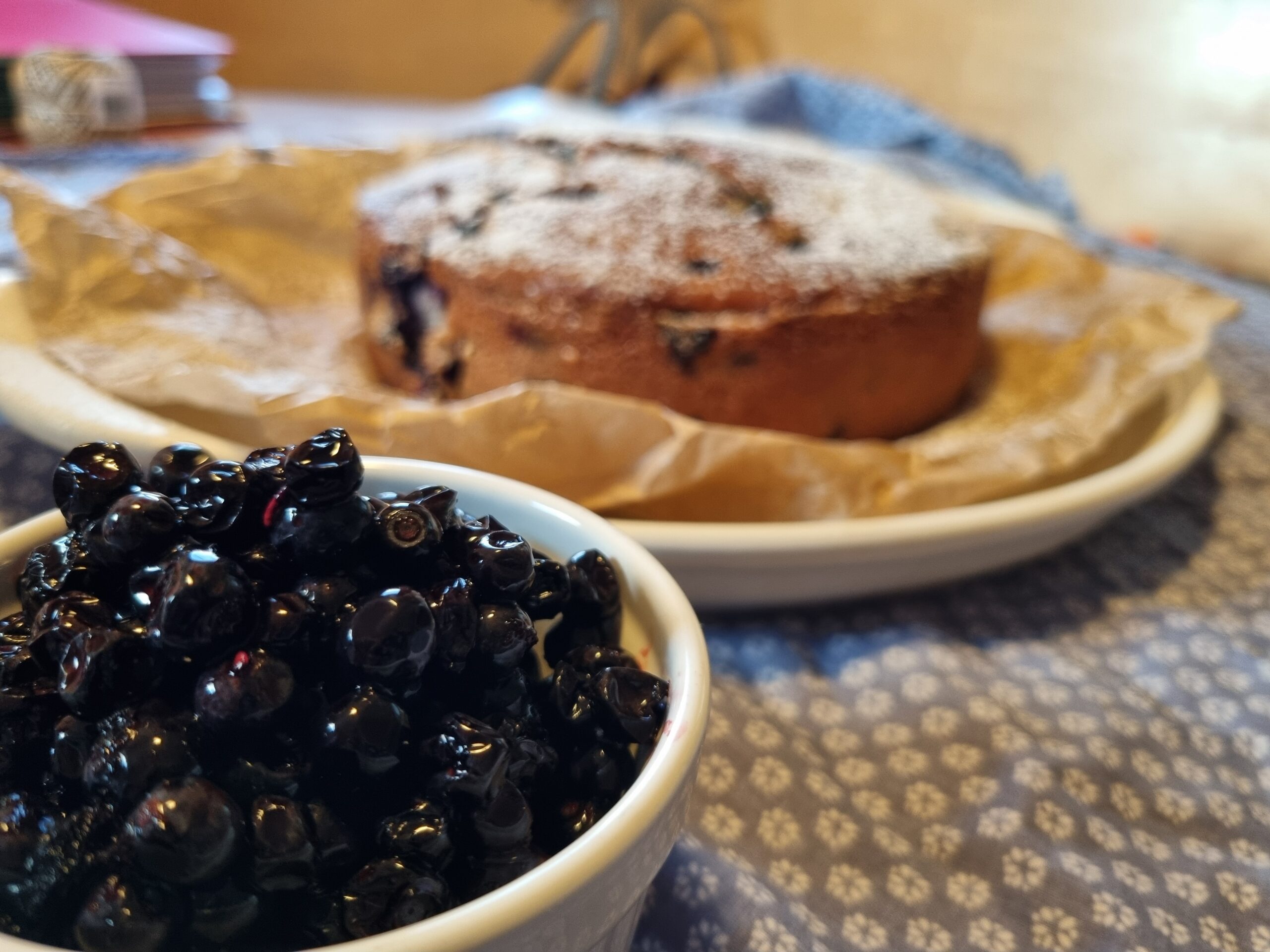 Eine Schale mit frischen Beeren vor einem mit Puderzucker bestäubten Kuchen auf einem Teller, der auf einem gemusterten blauen Tischtuch steht.