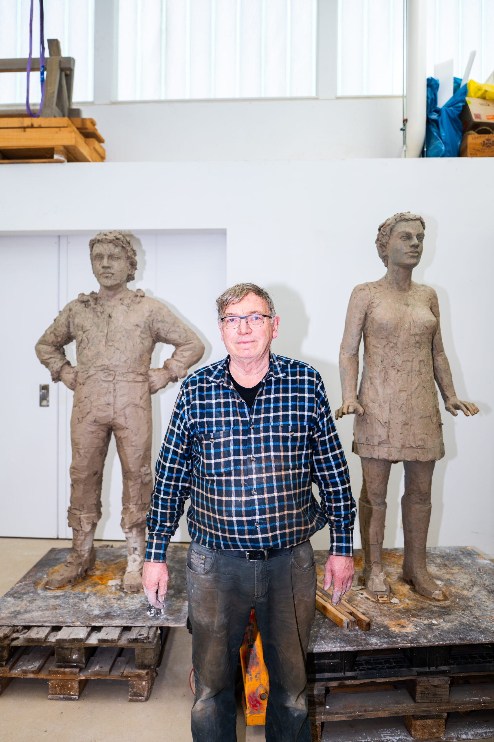 In einem hellen Atelier steht ein Mann vor zwei unfertigen Tonfiguren, einer m&auml;nnlichen und einer weiblichen.