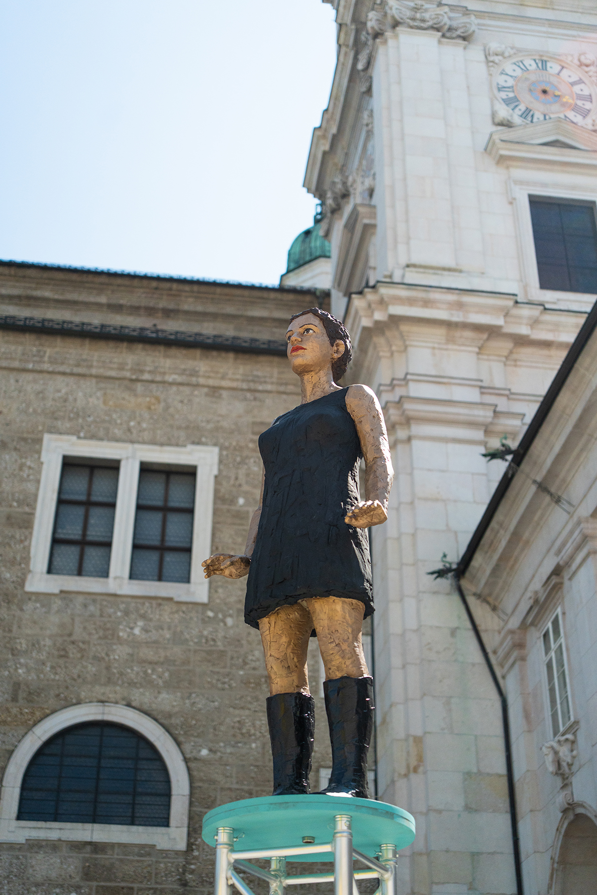 Auf einem Sockel vor einem historischen Geb&auml;ude steht eine Holzstatue einer Frau in einem schwarzen Kleid.