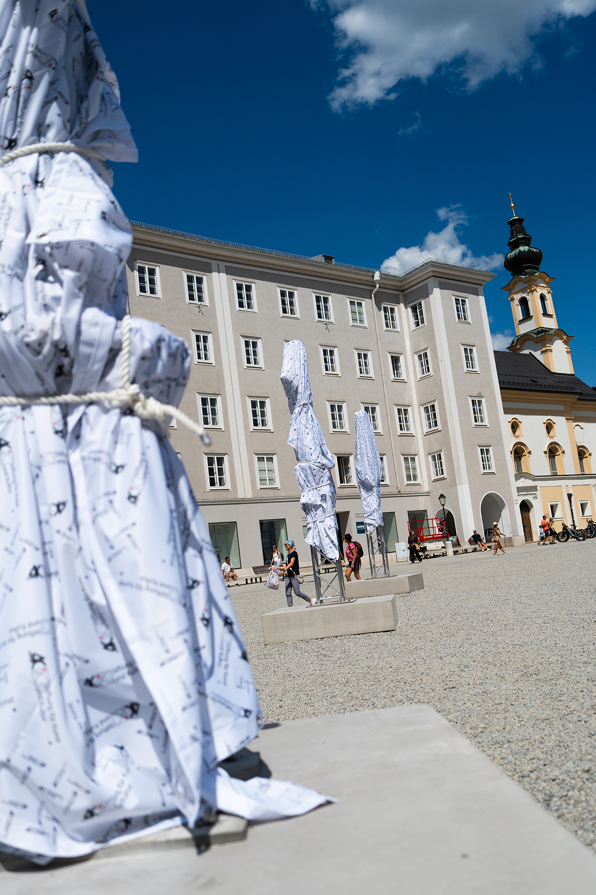 Ein Platz im Freien mit verh&uuml;llten Skulpturen, vorbeigehenden Menschen, einem beigen Geb&auml;ude und einer Kirche mit Uhrturm.