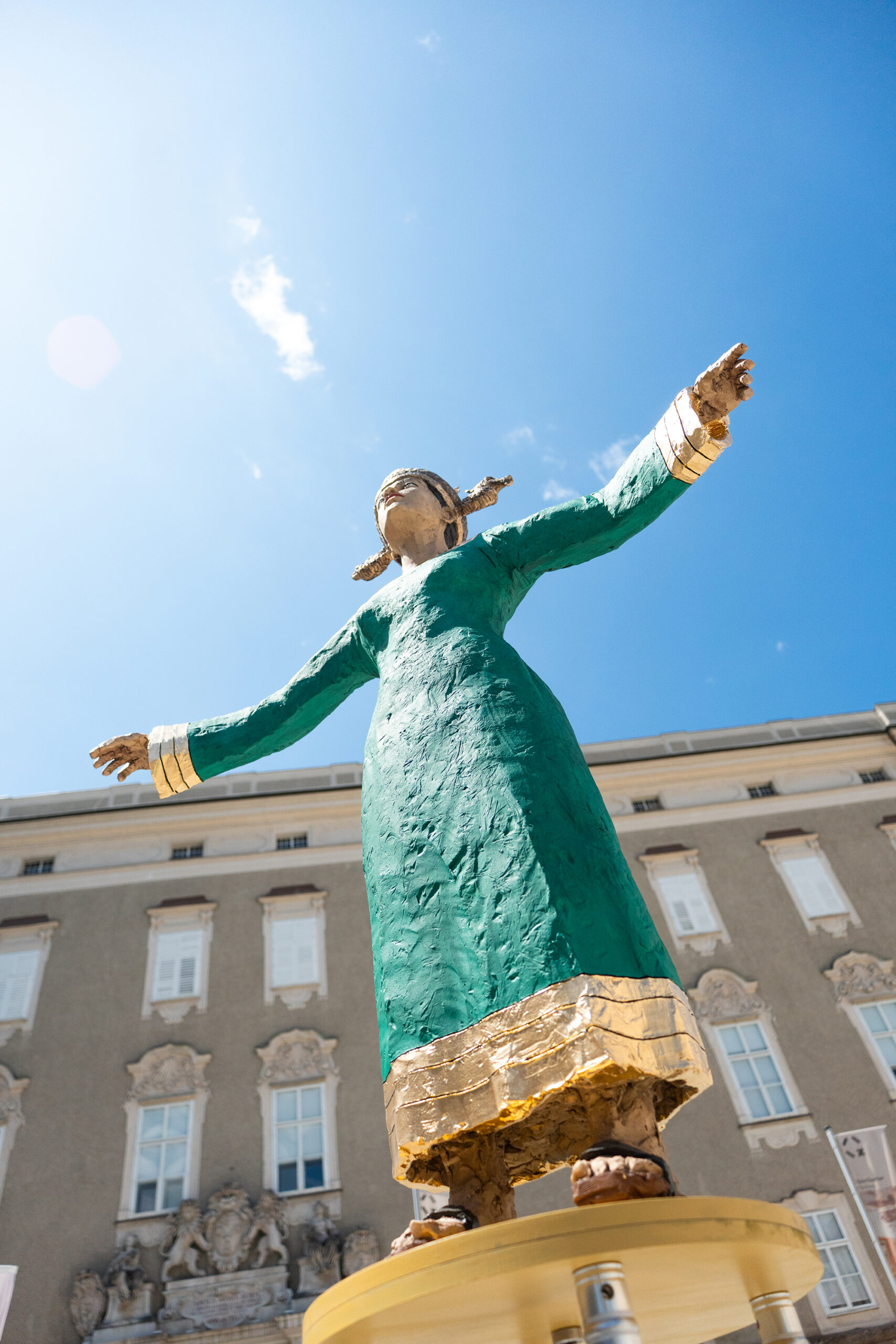 Gro&szlig;e Statue einer Frau in einem gr&uuml;nen Kleid mit ausgestreckten Armen, vor dem Hintergrund eines Geb&auml;udes und eines blauen Himmels.