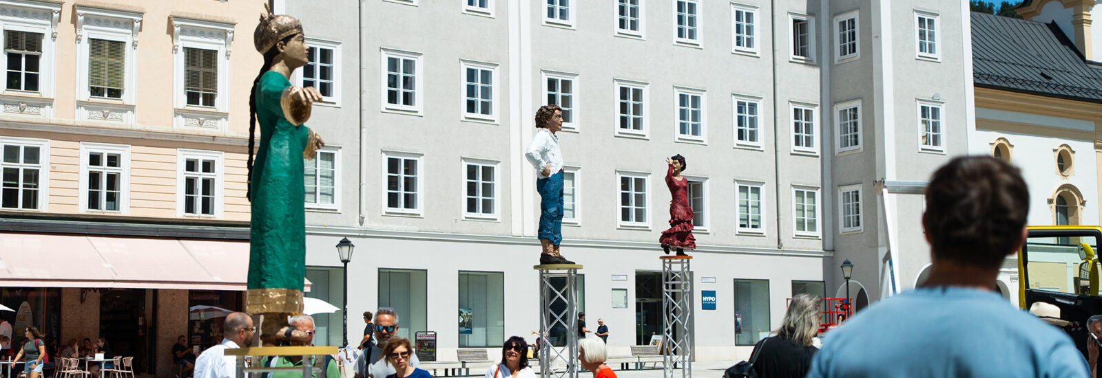 Auf einem europäischen Platz gehen Menschen vorbei, im Hintergrund stehen große menschliche Statuen auf Metallstativen und historische Gebäude.