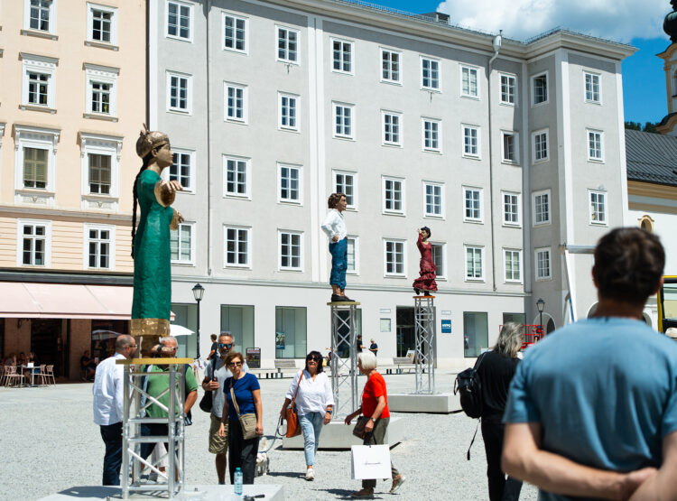 Auf einem europäischen Platz gehen Menschen vorbei, im Hintergrund stehen große menschliche Statuen auf Metallstativen und historische Gebäude.