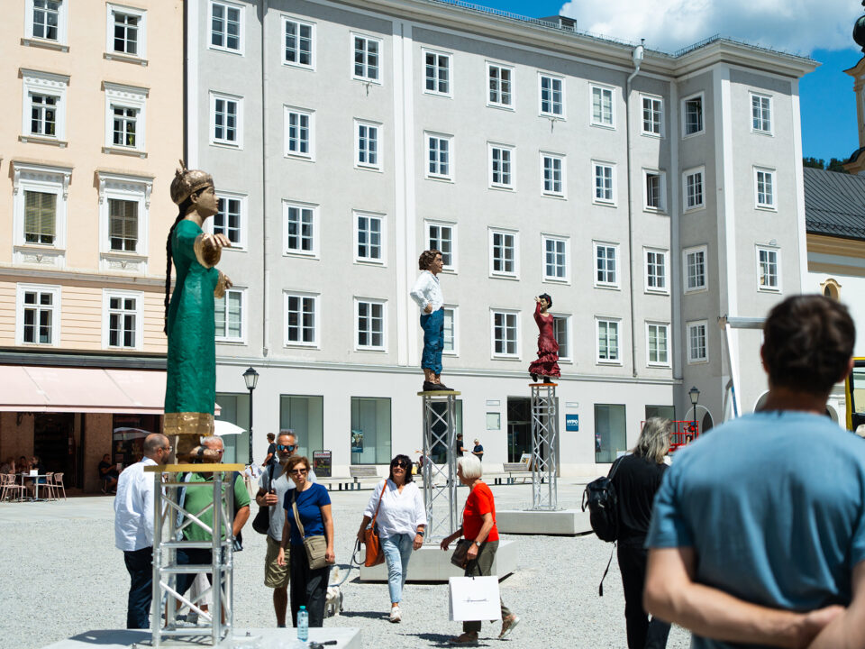 Auf einem europäischen Platz gehen Menschen vorbei, im Hintergrund stehen große menschliche Statuen auf Metallstativen und historische Gebäude.