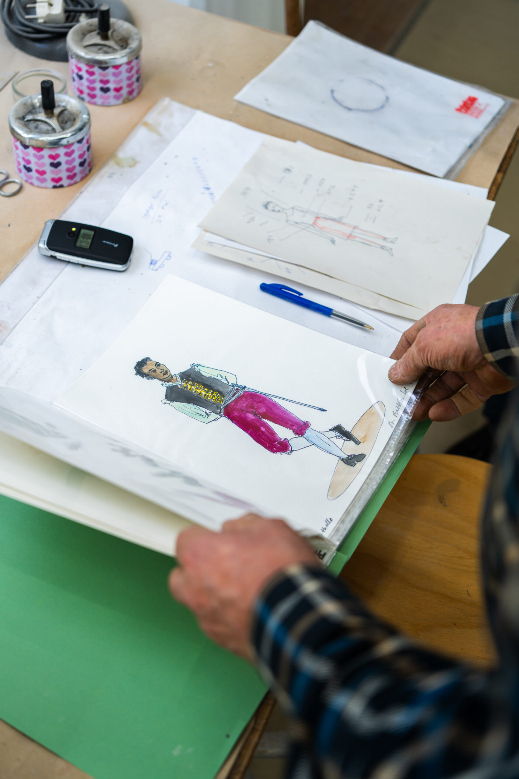 Eine Person betrachtet eine Modeskizze auf einem Schreibtisch, auf dem Zeichnungen, ein Telefon, Stifte und Klebeband liegen.