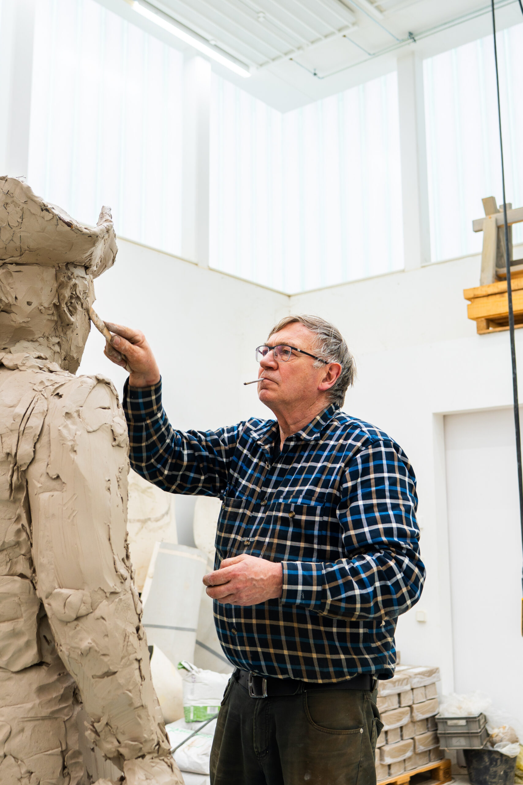 Ein &auml;lterer Mann, der in einem Raum eine gro&szlig;e Tonstatue modelliert; er tr&auml;gt eine Brille und ein kariertes Hemd.