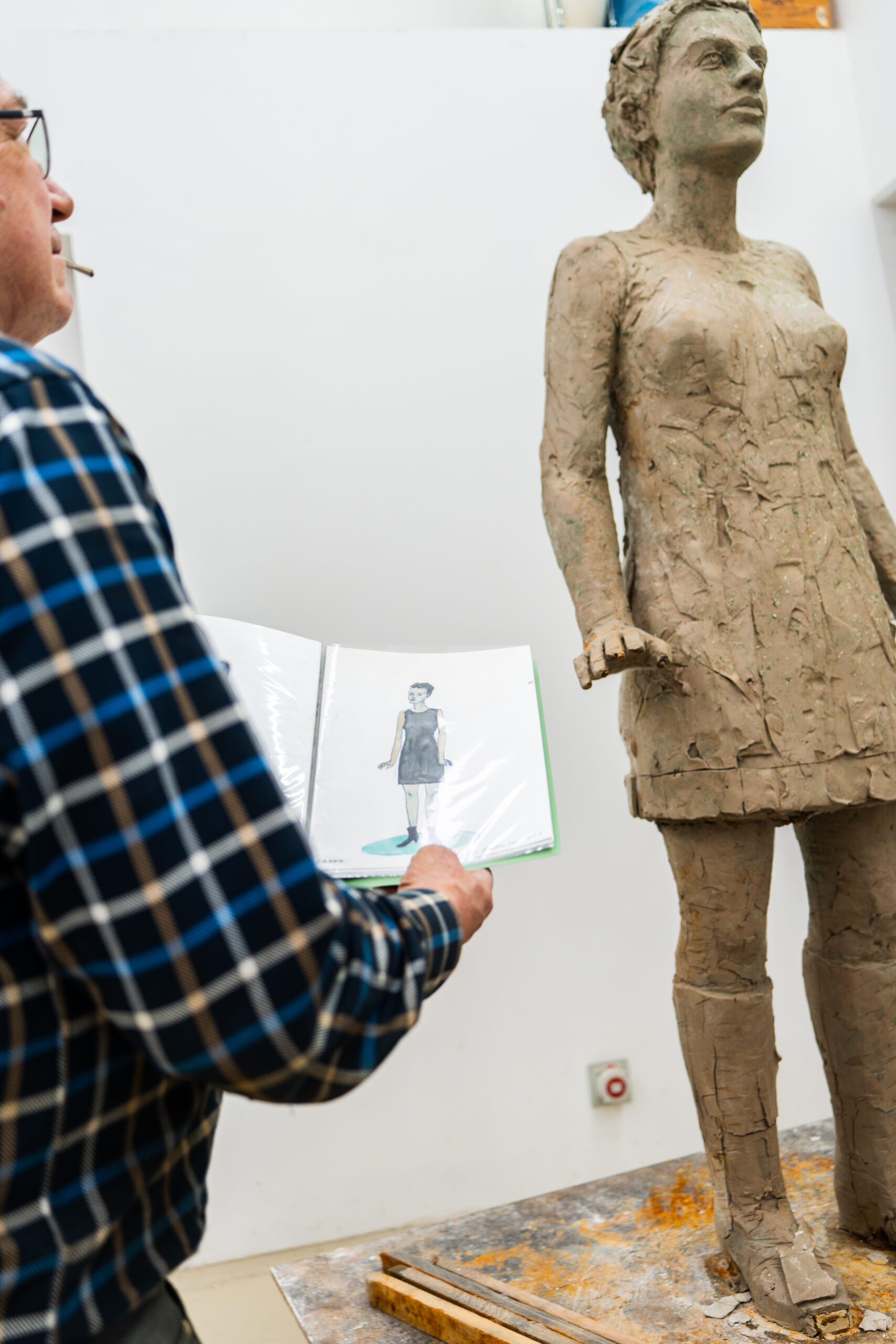 Eine Person h&auml;lt in einem Atelier eine Zeichnung neben einer Tonfigur einer Frau, die ein Kleid und Stiefel tr&auml;gt.