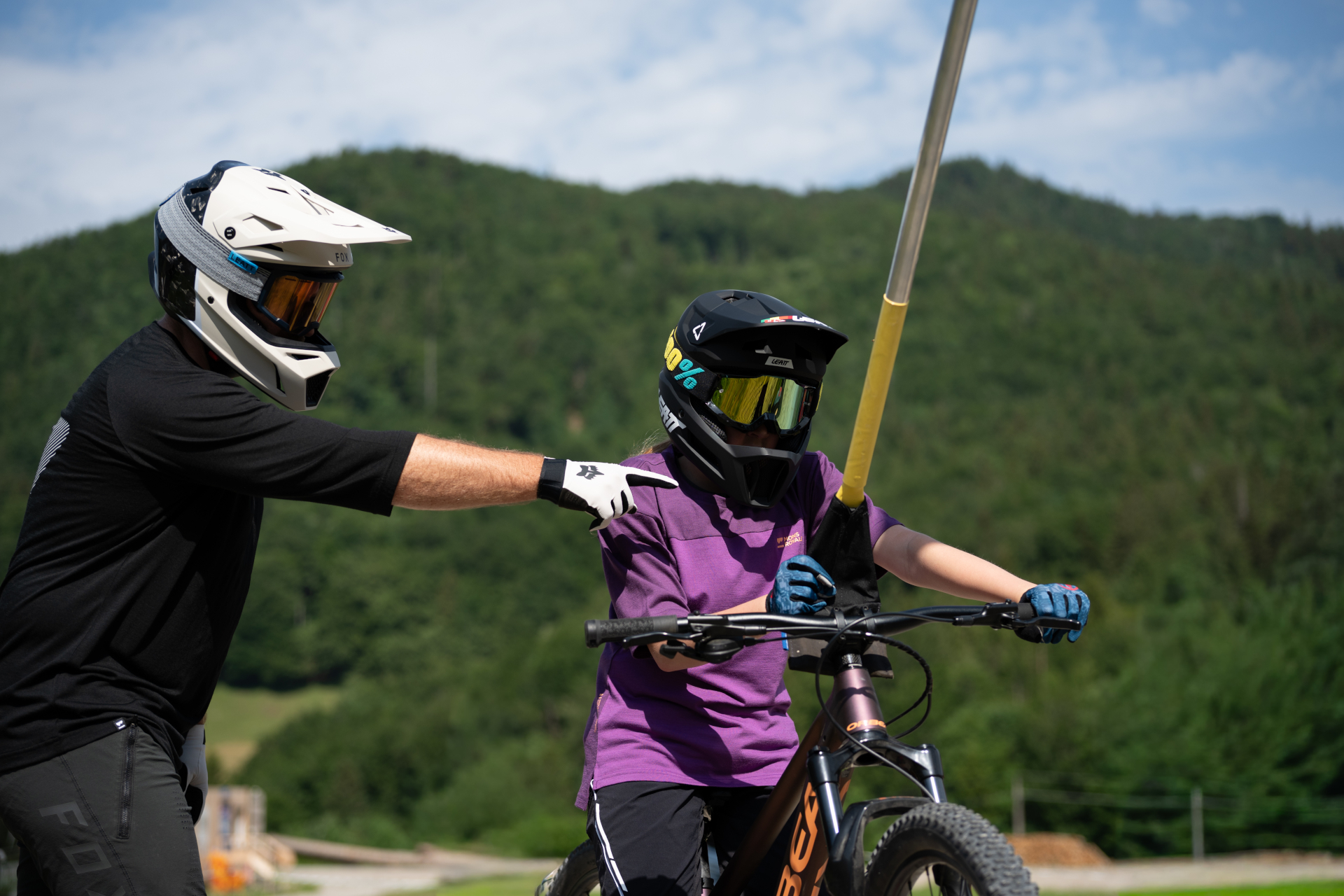Ein Erwachsener mit Helm, der ein Kind auf einem Mountainbike anleitet, im Hintergrund bewaldete Hügel.