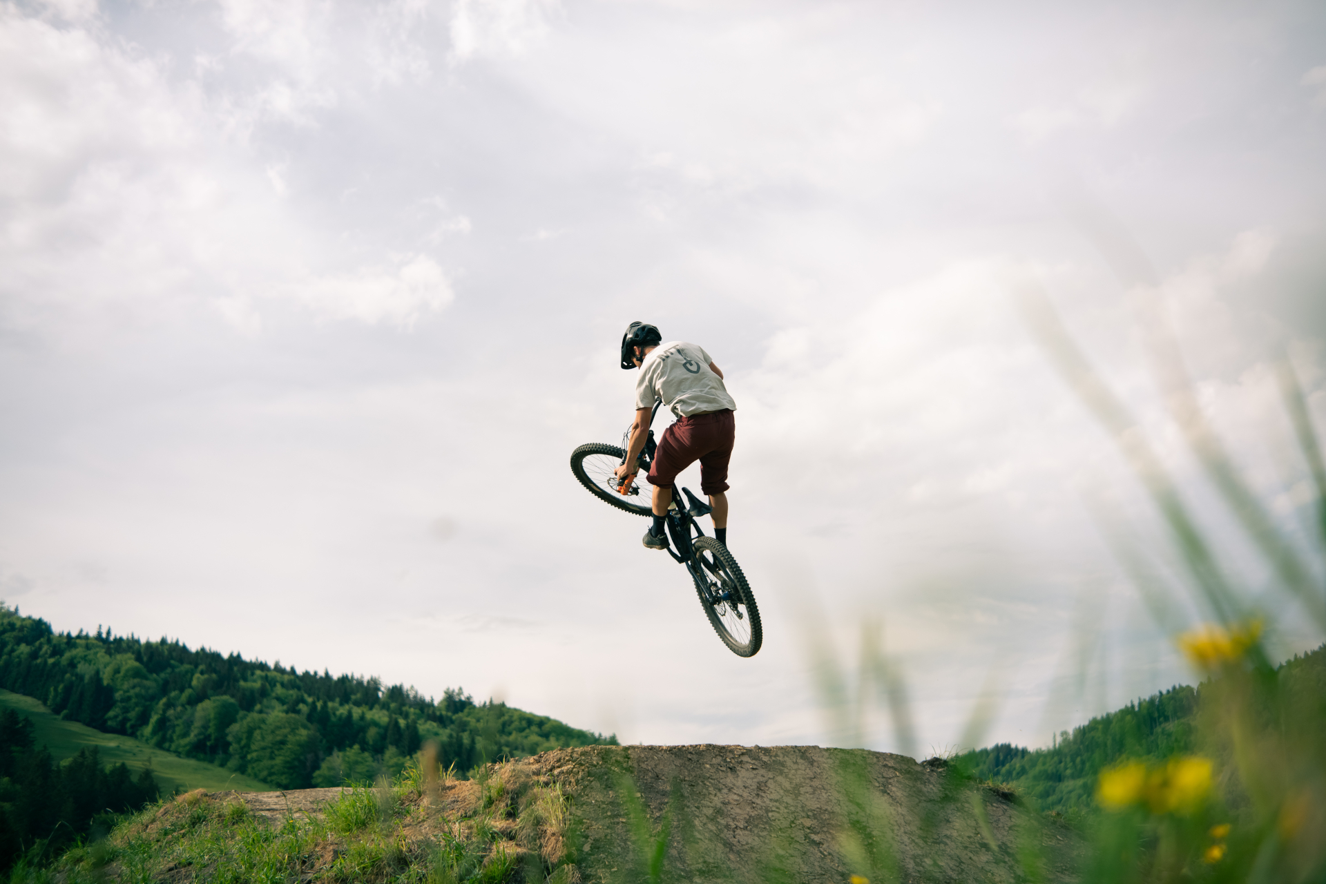 Eine Person mit Helm springt auf einem Mountainbike über einen Erdhügel, im Hintergrund sind Bäume zu sehen.