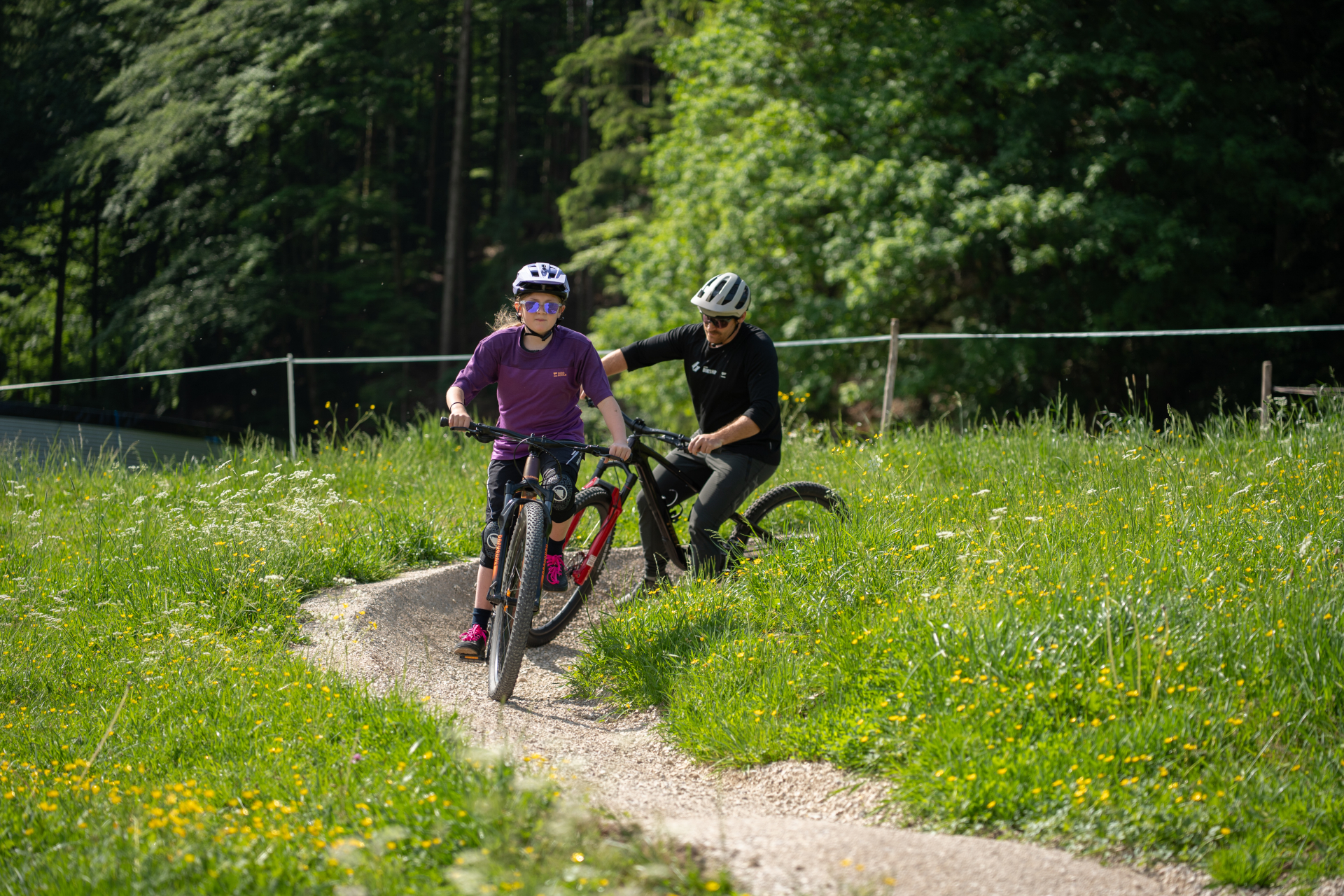 Zwei Personen mit Helmen fahren mit dem Fahrrad auf einem kurvenreichen Weg durch einen grasbewachsenen, sonnendurchfluteten Park, im Hintergrund sind Bäume zu sehen.