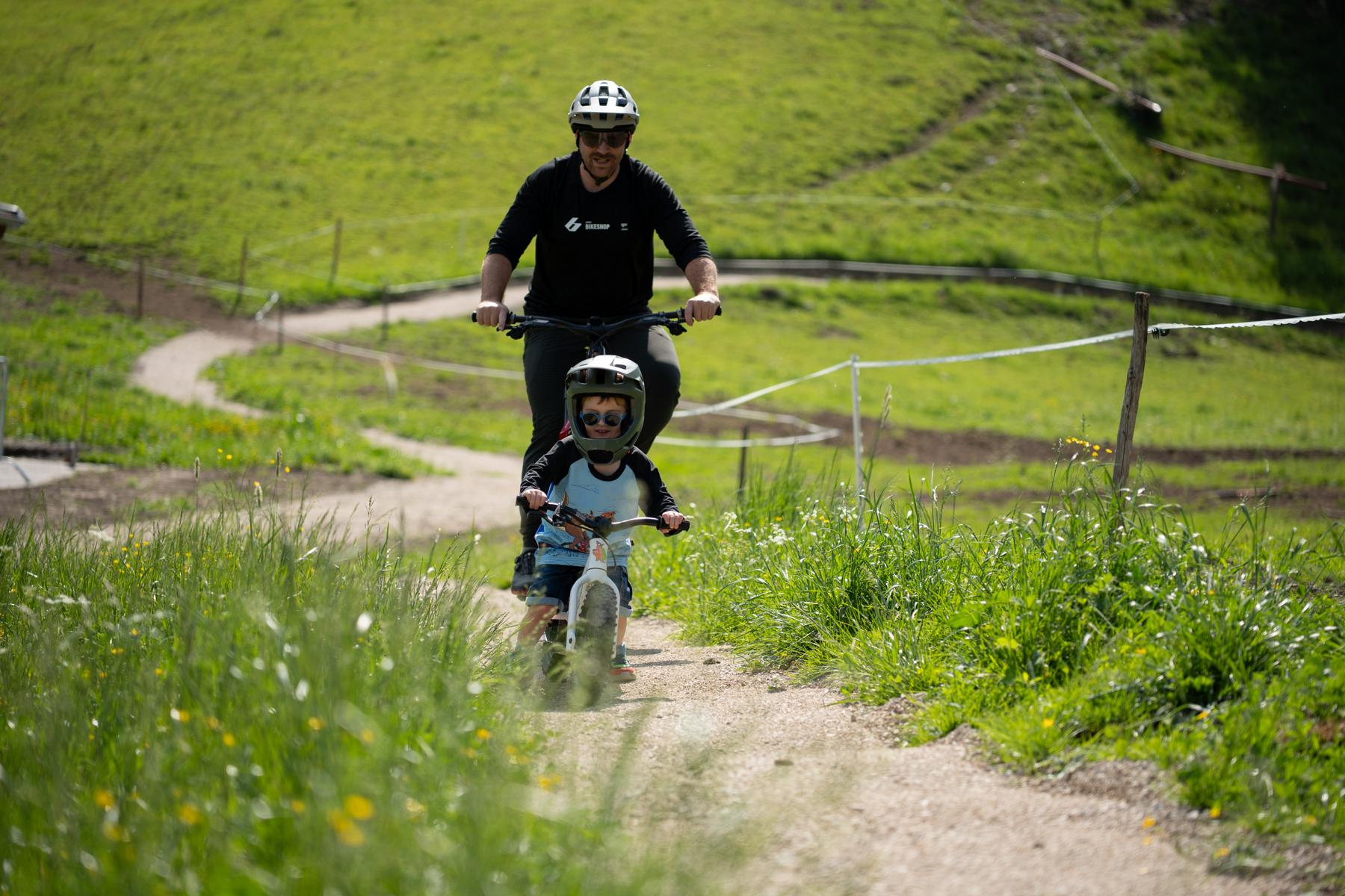 An einem sonnigen Tag fahren ein Erwachsener und ein Kind mit dem Fahrrad auf einem kurvenreichen Weg durch grasbewachsene Hügel; beide tragen einen Helm.