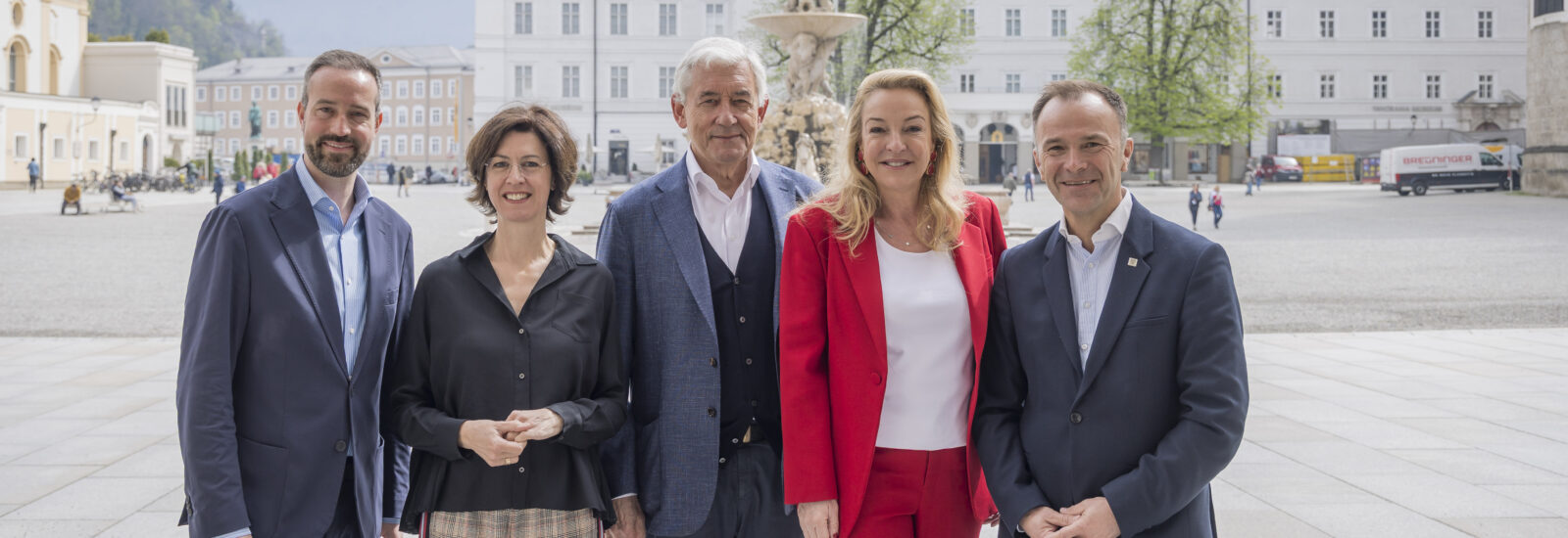 Fünf Personen in Geschäftskleidung posieren gemeinsam im Freien auf einem europäischen Stadtplatz, hinter ihnen sind Gebäude zu sehen.