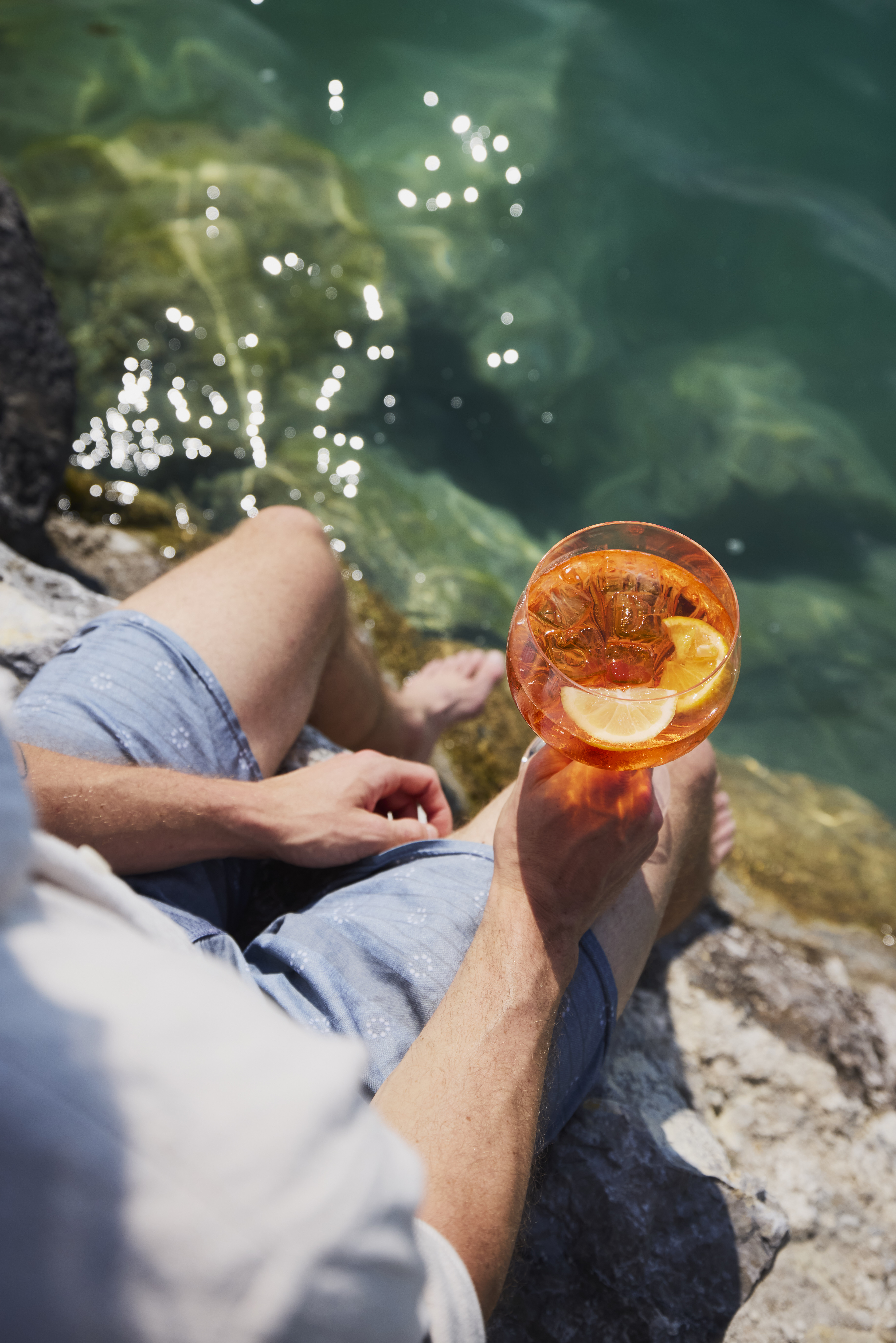 Eine Person sitzt am klaren Wasser, h&auml;lt einen orangefarbenen Cocktail mit einer Zitronenscheibe in der Hand und hat die F&uuml;&szlig;e nahe am Ufer.