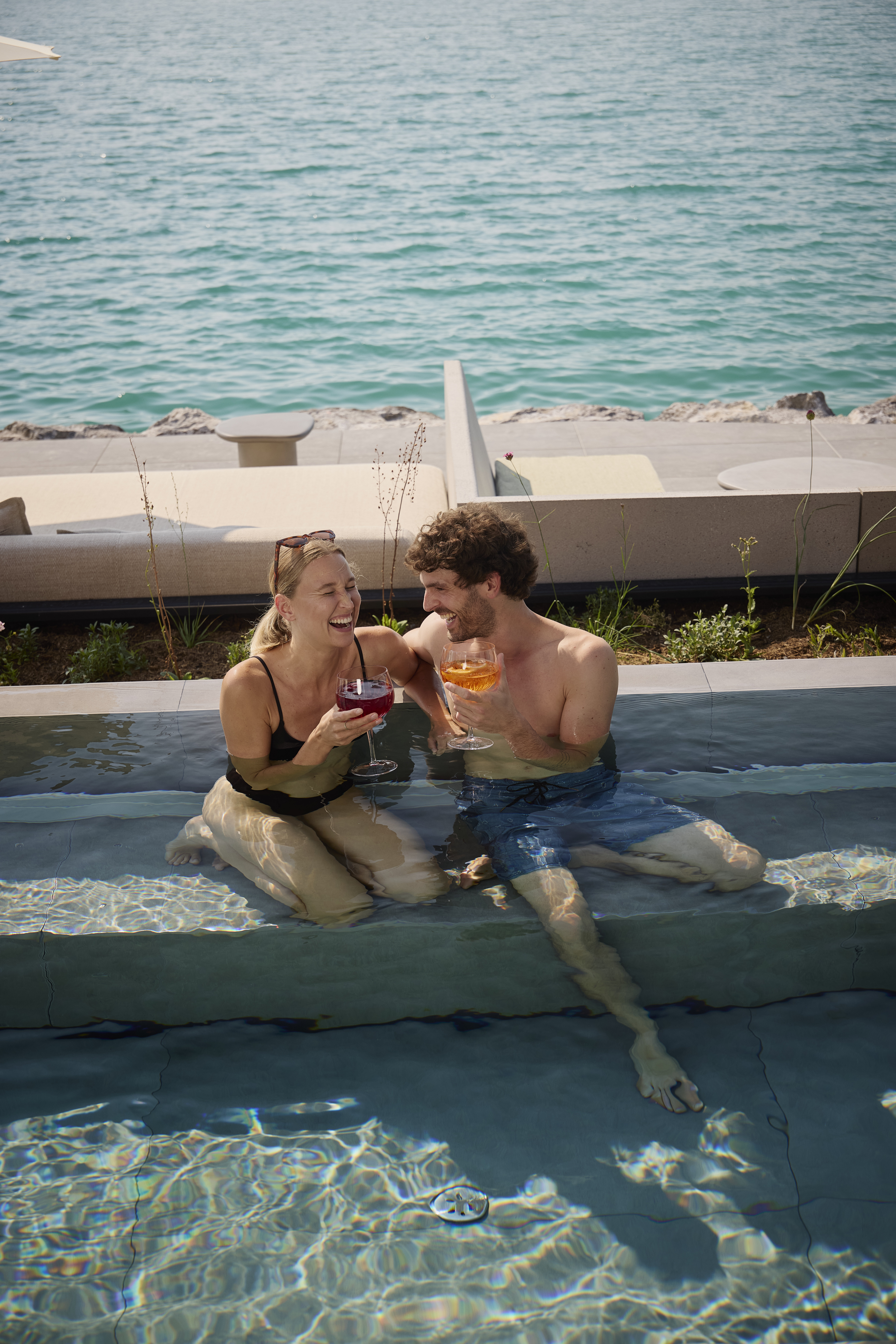 Ein l&auml;chelndes Paar in Badekleidung genie&szlig;t gemeinsam einen Drink, w&auml;hrend es in einem Pool am Meer sitzt.