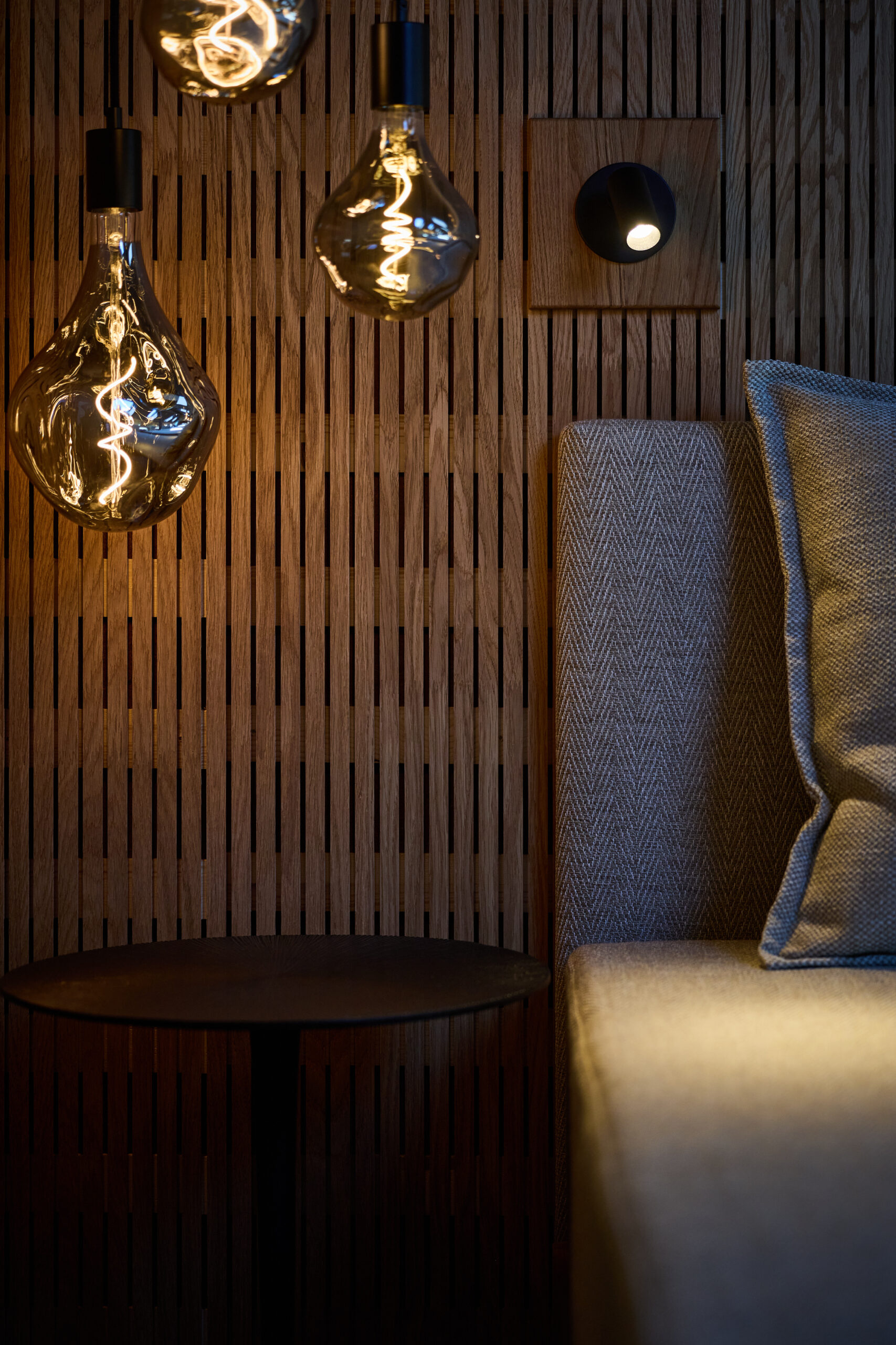 Modernes, gem&uuml;tliches Interieur mit h&auml;ngenden Edison-Gl&uuml;hbirnen, einer Wand aus Holzlatten, einem Beistelltisch und einem Sofa mit Kissen.