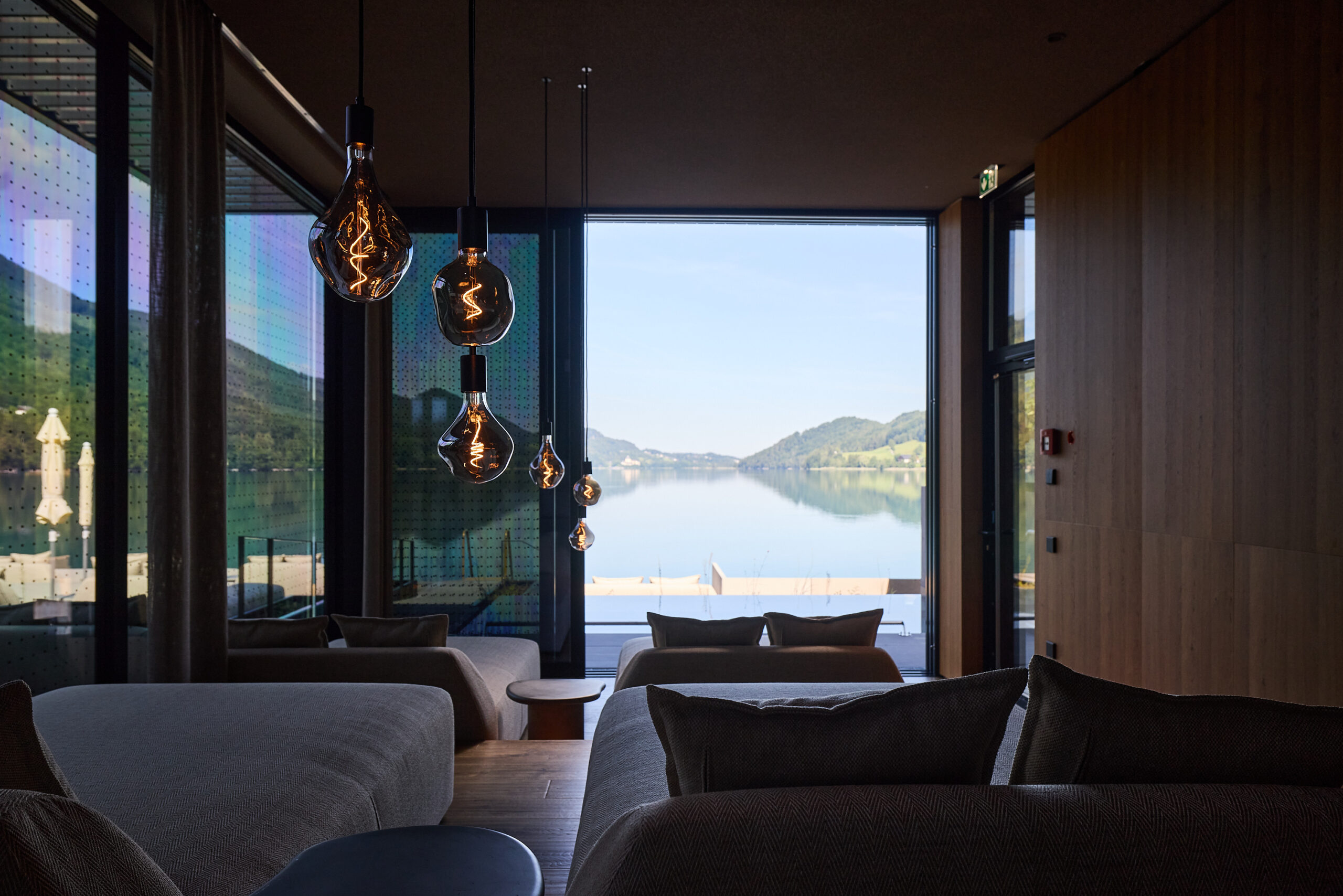 Moderne Lounge mit H&auml;ngeleuchten, gro&szlig;en Fenstern und einem malerischen Blick auf den See und die Berge.