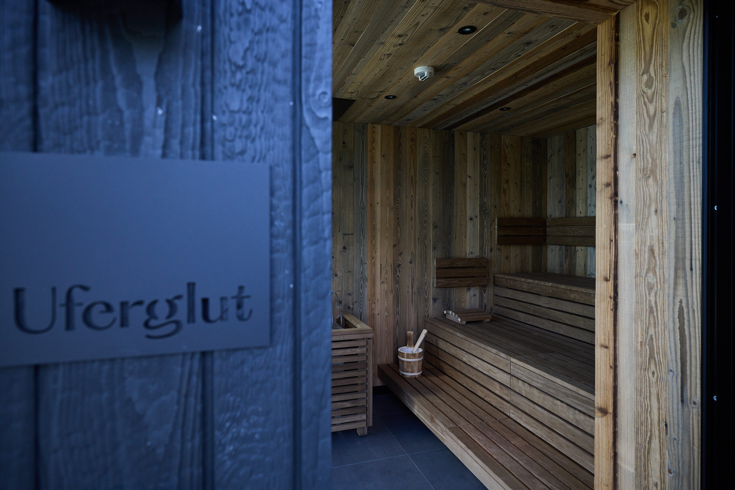 Sauna-Innenraum mit Holzvert&auml;felung, B&auml;nken und einem Eimer; &bdquo;Uferglut&ldquo;-Schild an der dunklen Au&szlig;enwand.