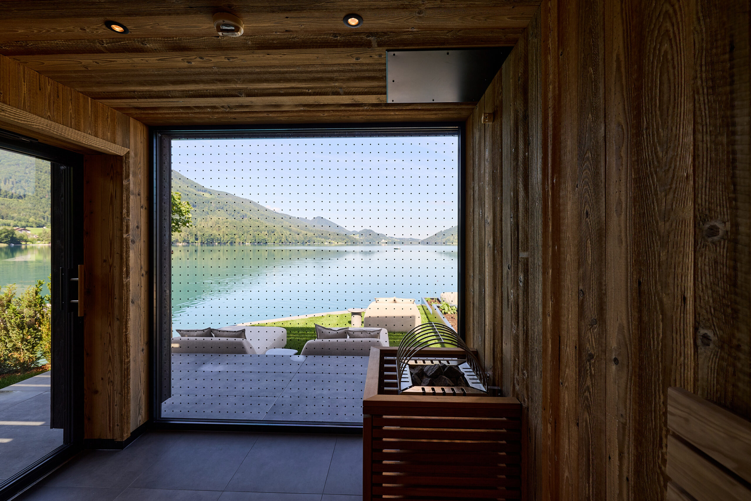 Blick durch eine Holzsauna auf einen See, die Berge und eine Terrasse mit Liegest&uuml;hlen an einem sonnigen Tag.