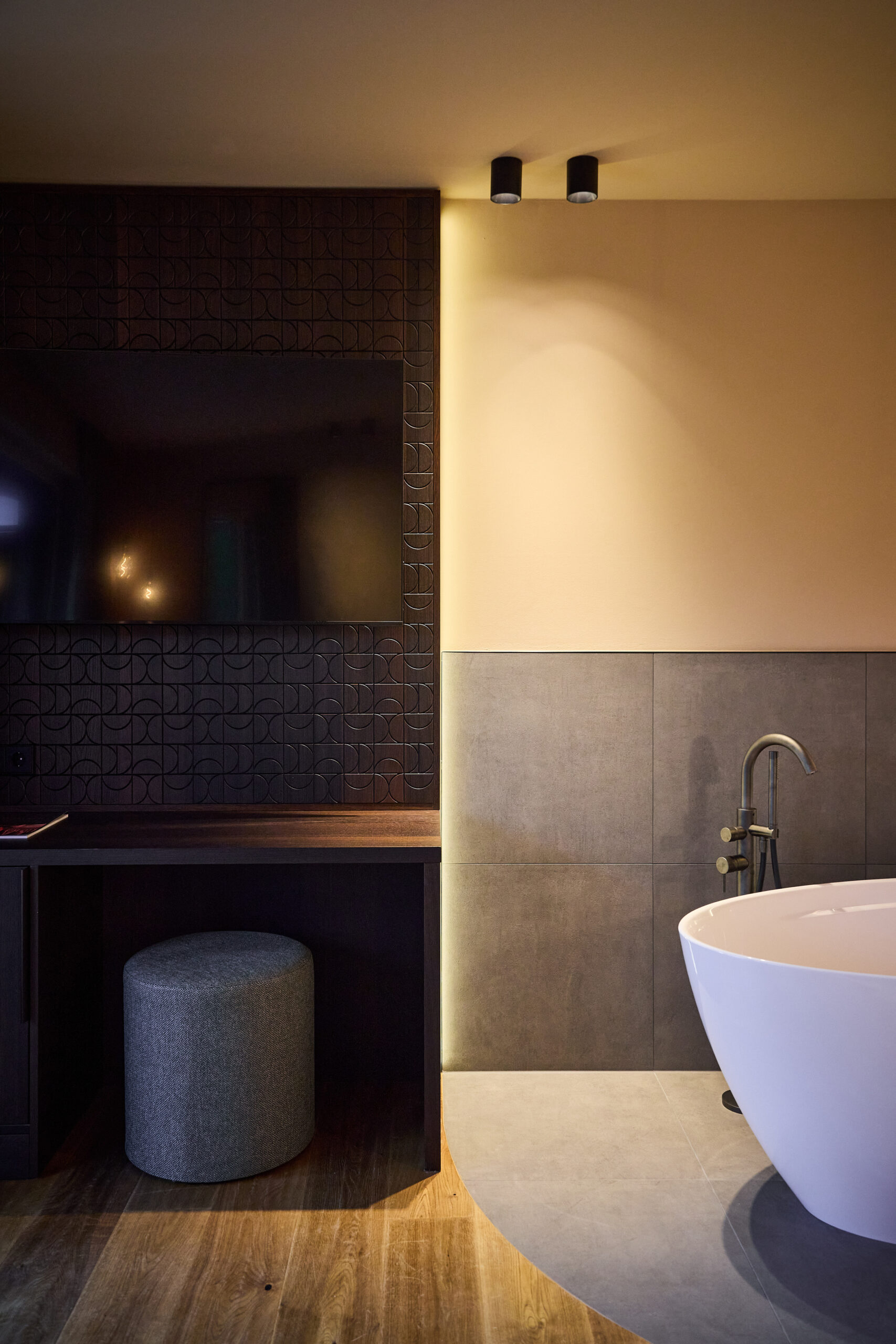 Modernes Badezimmer mit einer freistehenden Badewanne, einer wandmontierten Armatur, einem Schreibtischbereich und einem Fernseher vor einer gemusterten Wand.