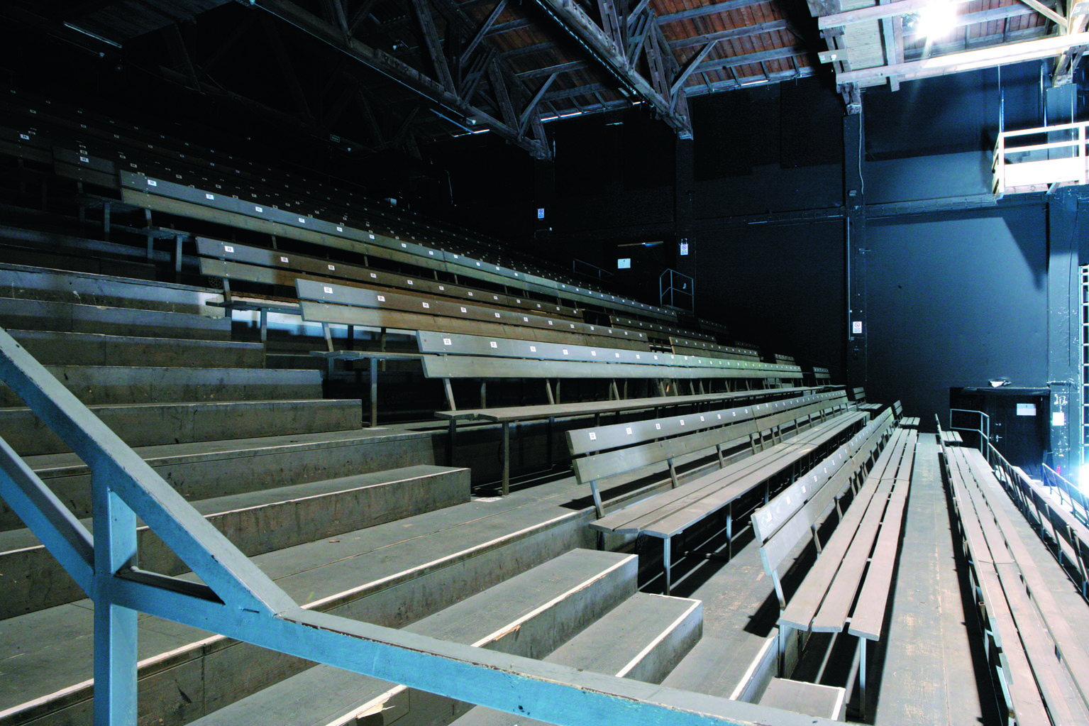Leere Holzsitze auf einer Trib&uuml;ne in einer Halle mit Metallgel&auml;ndern und freiliegenden Balken &uuml;ber den Sitzen.