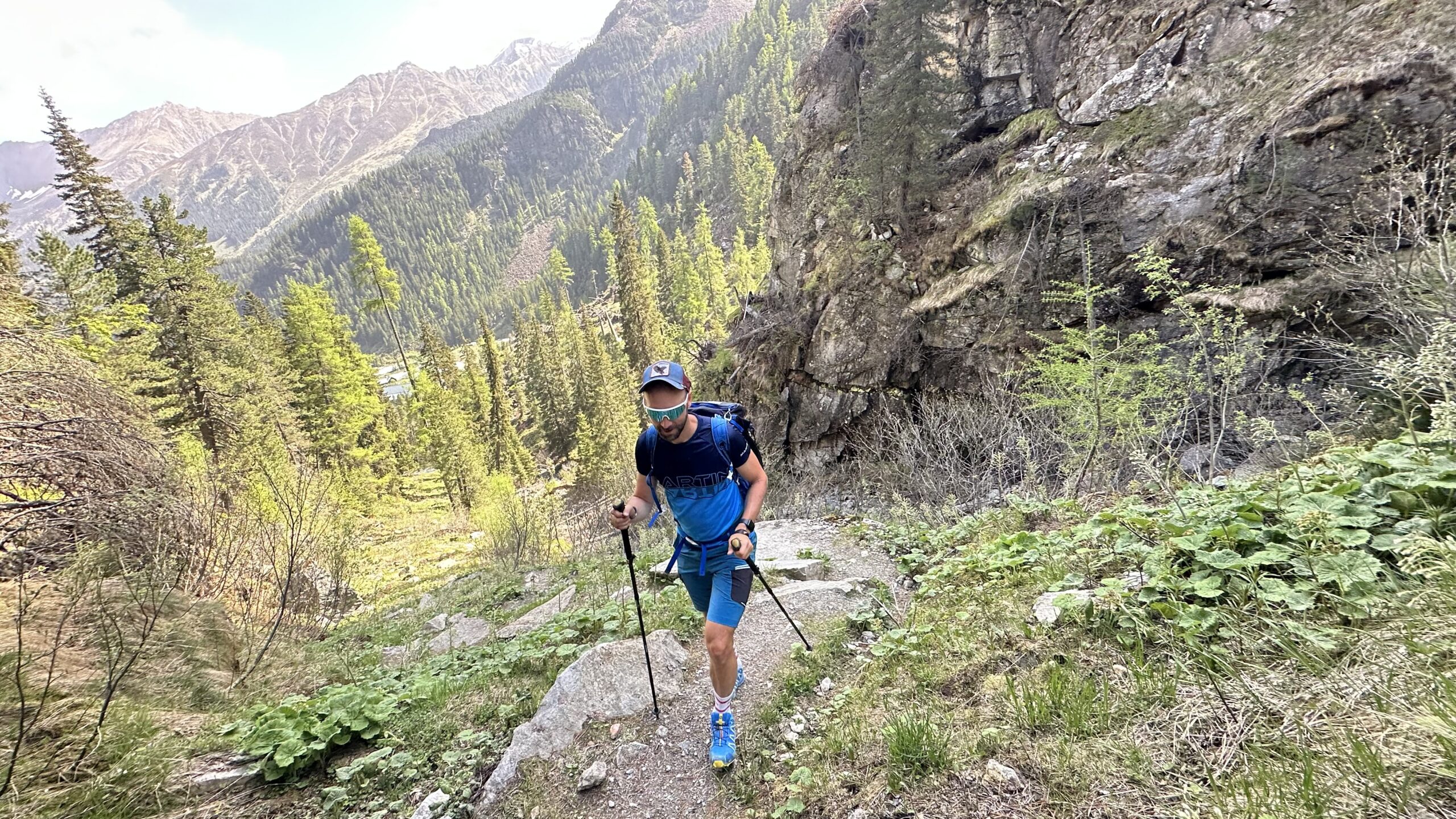 Eine Person mit Wanderst&ouml;cken geht einen felsigen Bergpfad hinauf, der von B&auml;umen und gr&uuml;nem Laub umgeben ist.