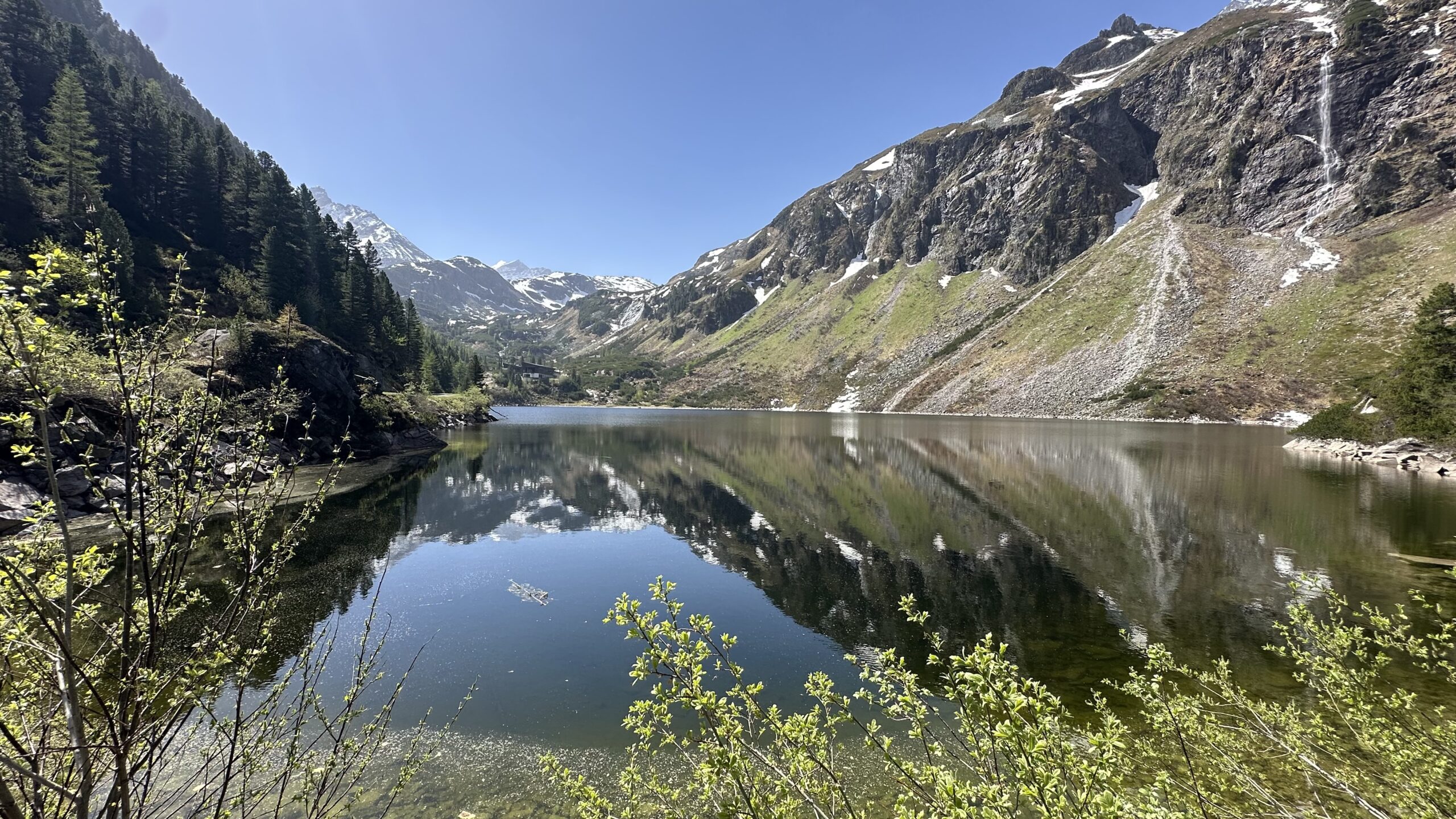 Ein klarer See spiegelt schneebedeckte Berge und B&auml;ume unter einem strahlend blauen Himmel wider.