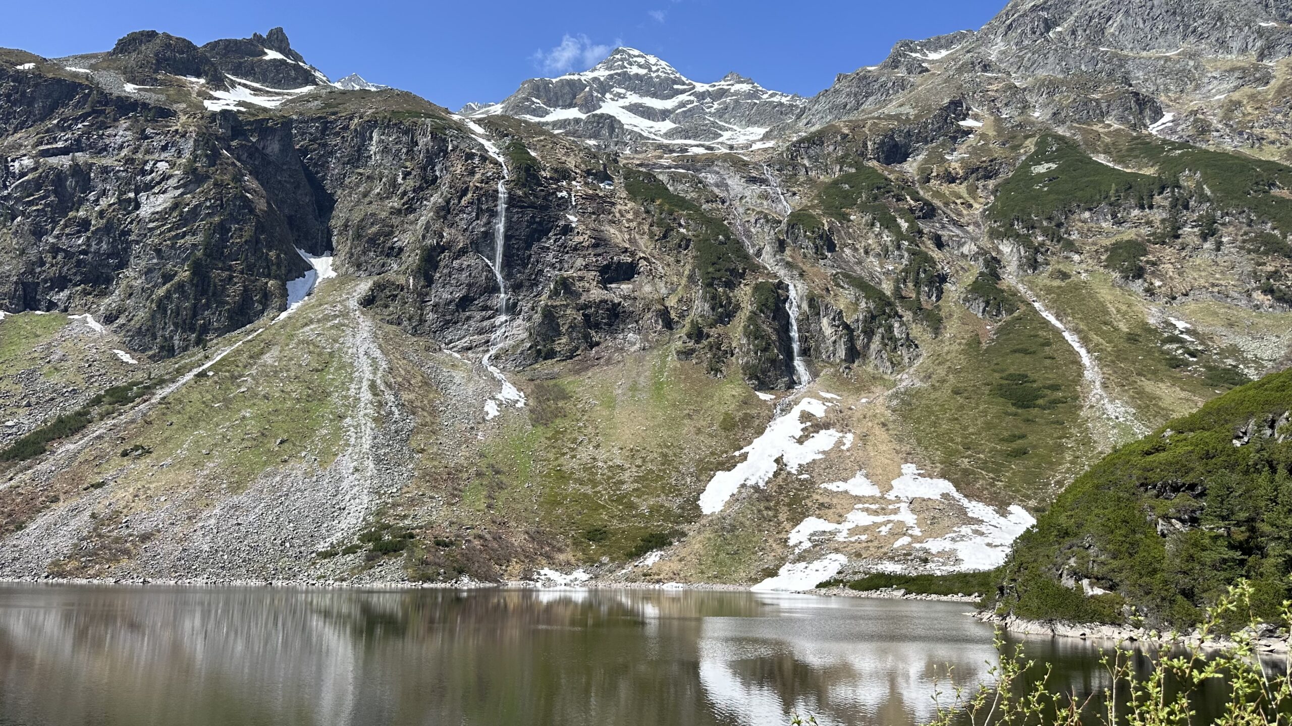 Schneebedeckte Berge mit felsigen H&auml;ngen spiegeln sich in einem ruhigen See unter einem strahlend blauen Himmel.