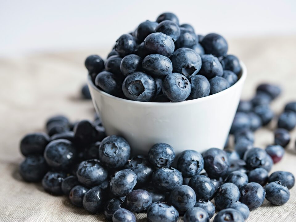 Eine weiße Schüssel, gefüllt mit frischen Heidelbeeren, um die herum weitere Heidelbeeren auf einer beigen Fläche verstreut liegen.