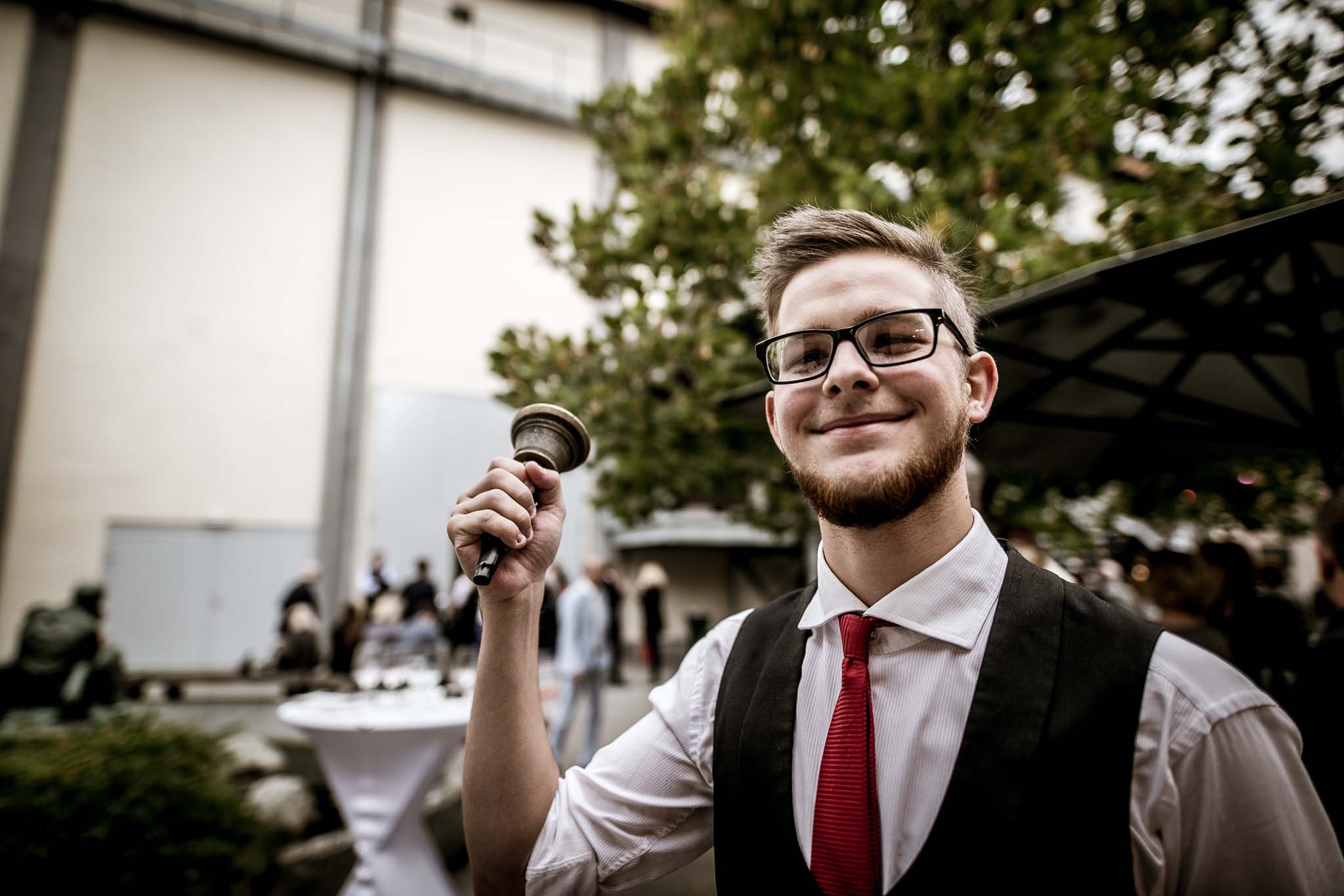 Ein l&auml;chelnder Mann mit Brille und roter Krawatte l&auml;utet bei einer Veranstaltung im Freien eine Glocke, w&auml;hrend im Hintergrund Menschen zu sehen sind.
