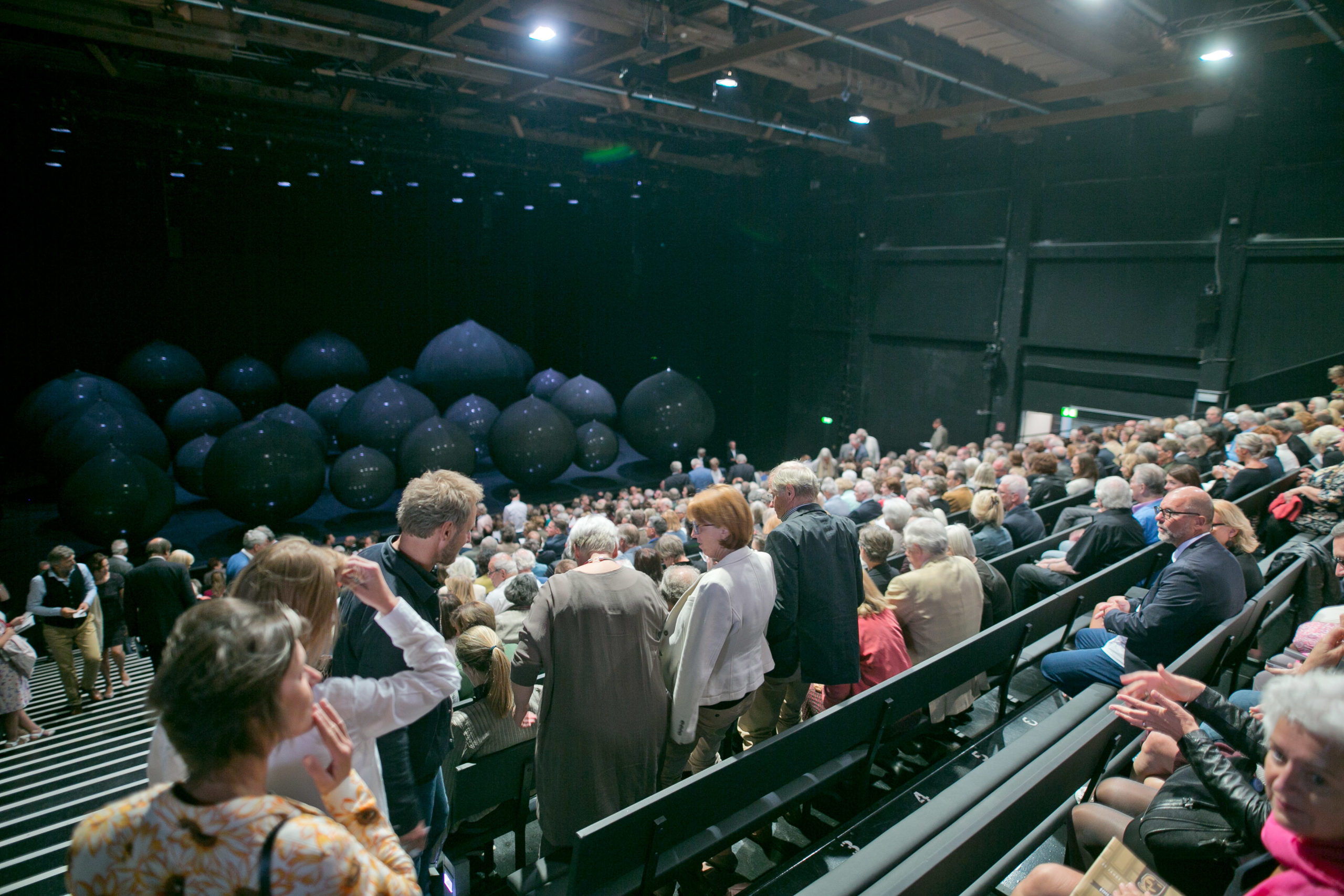 Vor Beginn einer Vorstellung f&uuml;llt eine Menschenmenge die Theaterpl&auml;tze, w&auml;hrend gro&szlig;e schwarze Luftballons auf der B&uuml;hne liegen.