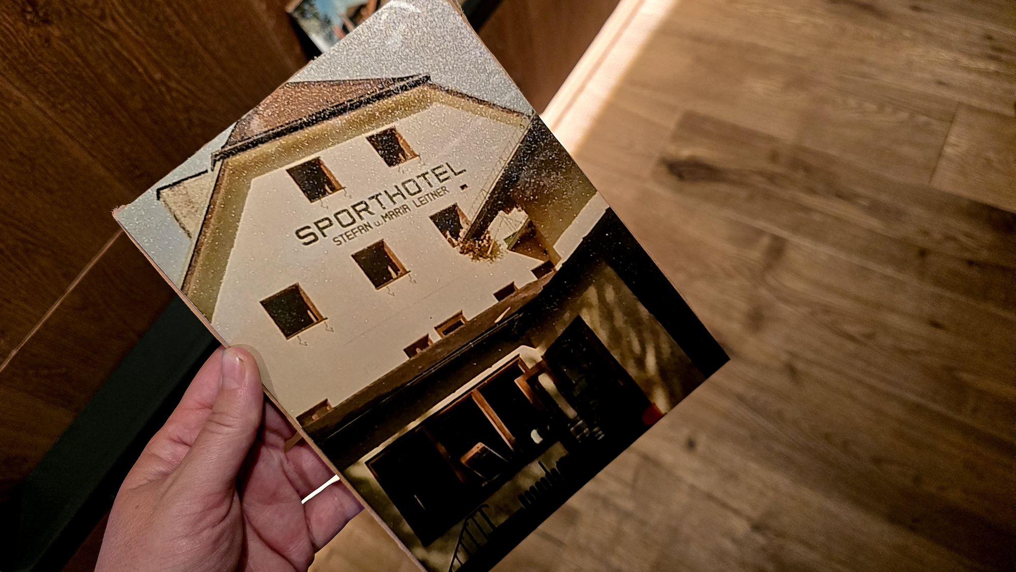 Eine Hand h&auml;lt ein Foto eines Geb&auml;udes mit der Aufschrift &bdquo;SPORTHOTEL&ldquo; &uuml;ber einem Holzboden.