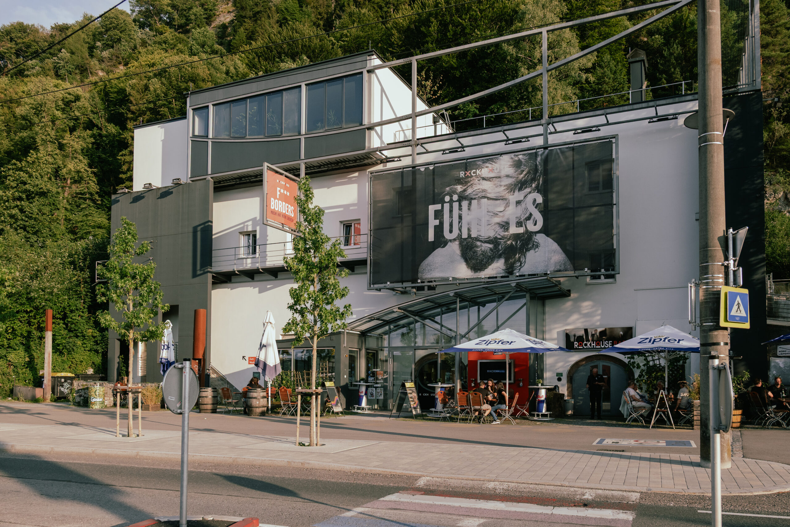 Modernes Geb&auml;ude mit einem gro&szlig;en Schwarz-Wei&szlig;-Filmplakat, einem B&uuml;rgersteig, B&auml;umen und einer Sitzgelegenheit im Freien.