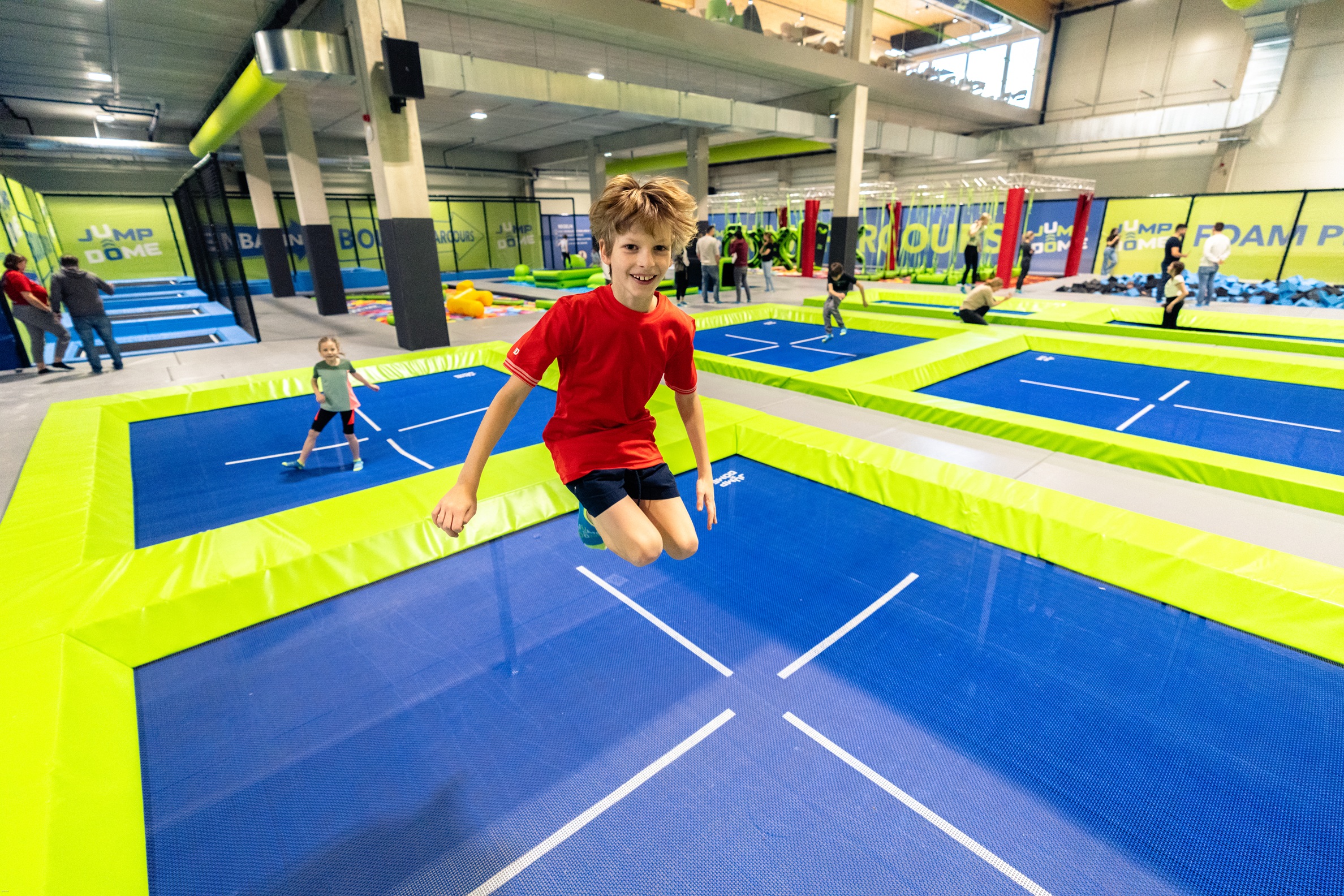 Ein Junge in einem roten T-Shirt springt auf einem Trampolin in einem Indoor-Park, im Hintergrund sind andere Kinder zu sehen.