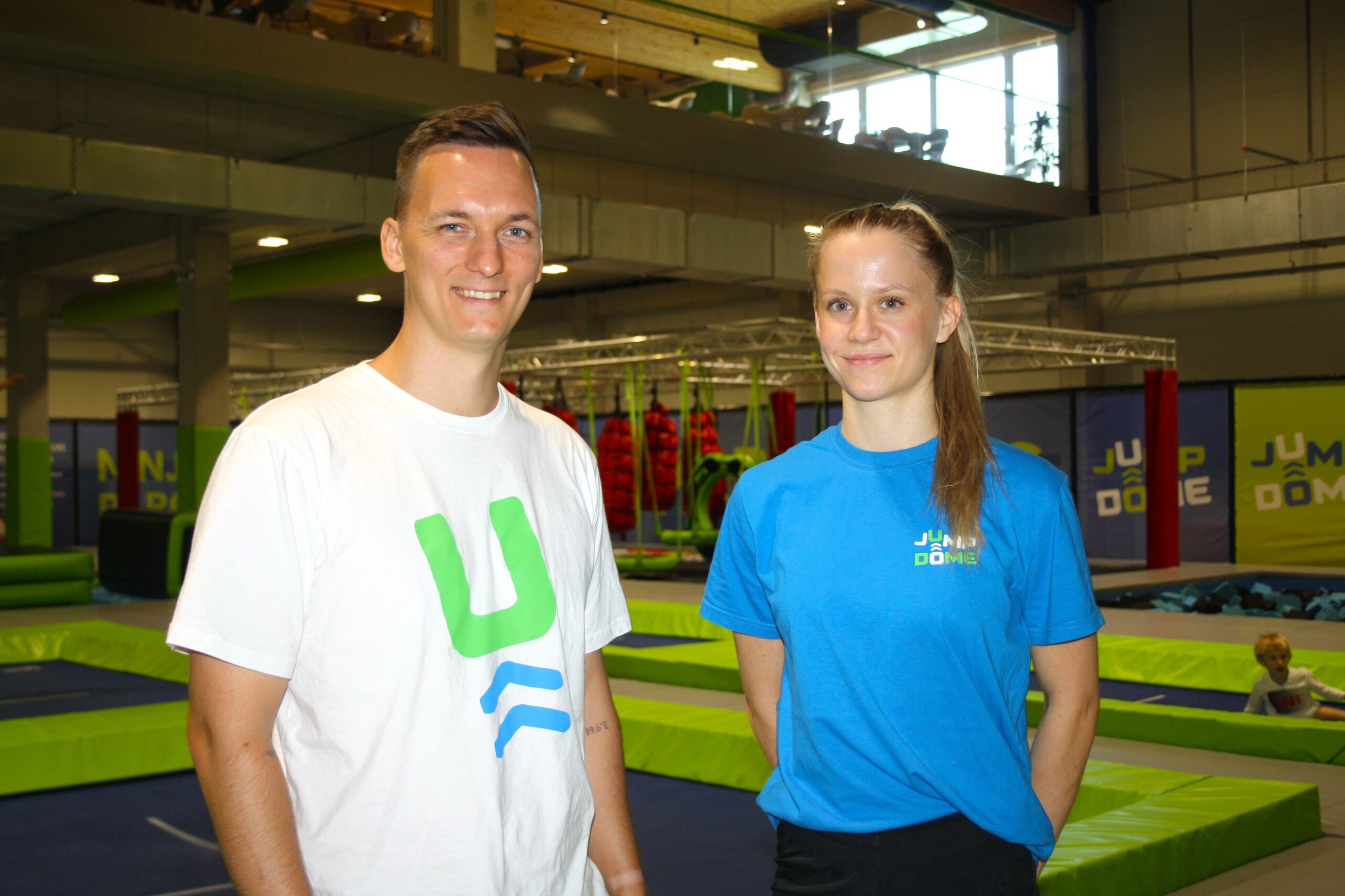 Zwei Personen in Sportshirts l&auml;cheln in einem Indoor-Trampolinpark mit leuchtend gr&uuml;nen und blauen Ger&auml;ten.