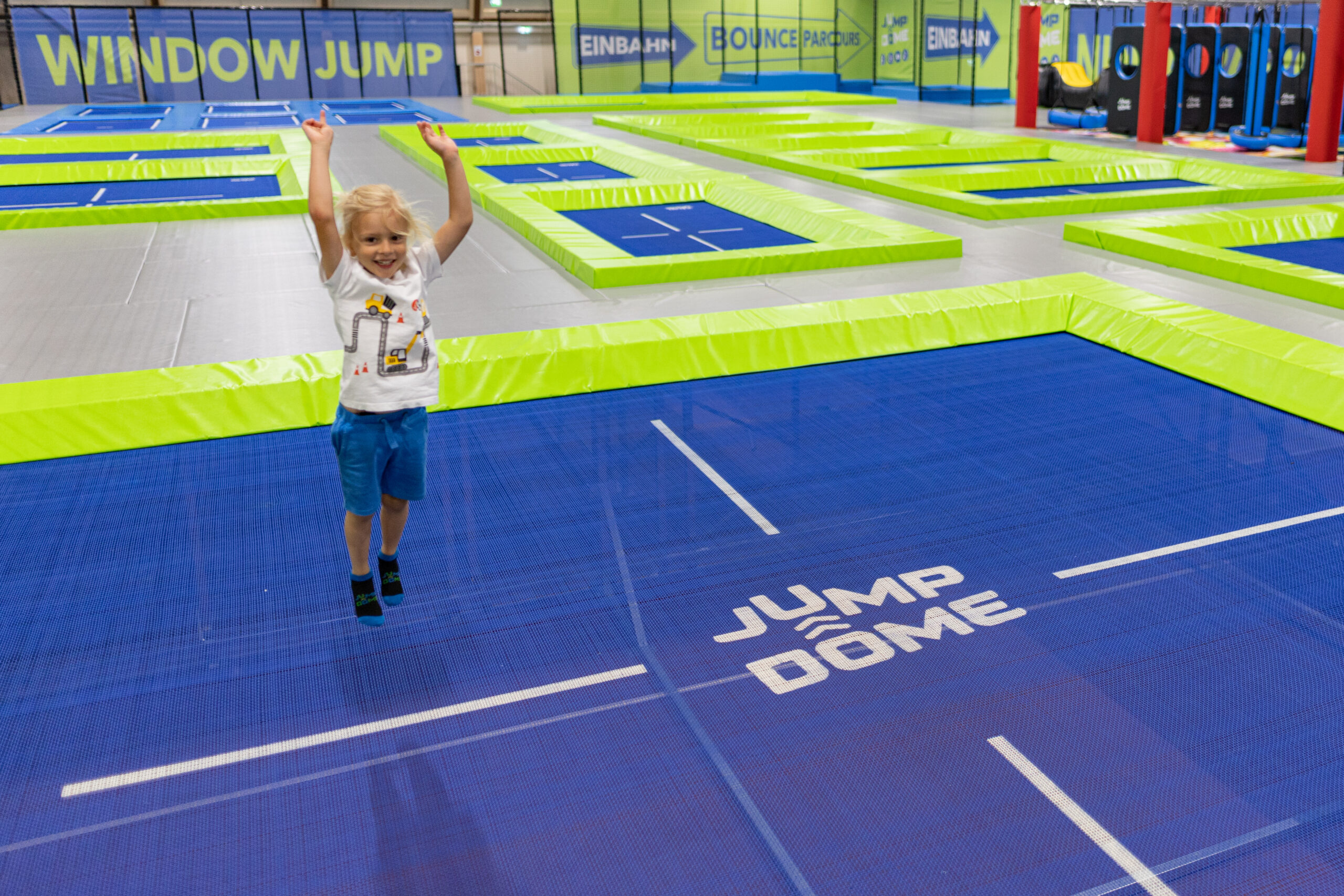 Ein l&auml;chelndes Kind, das mit erhobenen Armen auf einem blau-gr&uuml;nen Trampolin in einem Indoor-Park herumh&uuml;pft.