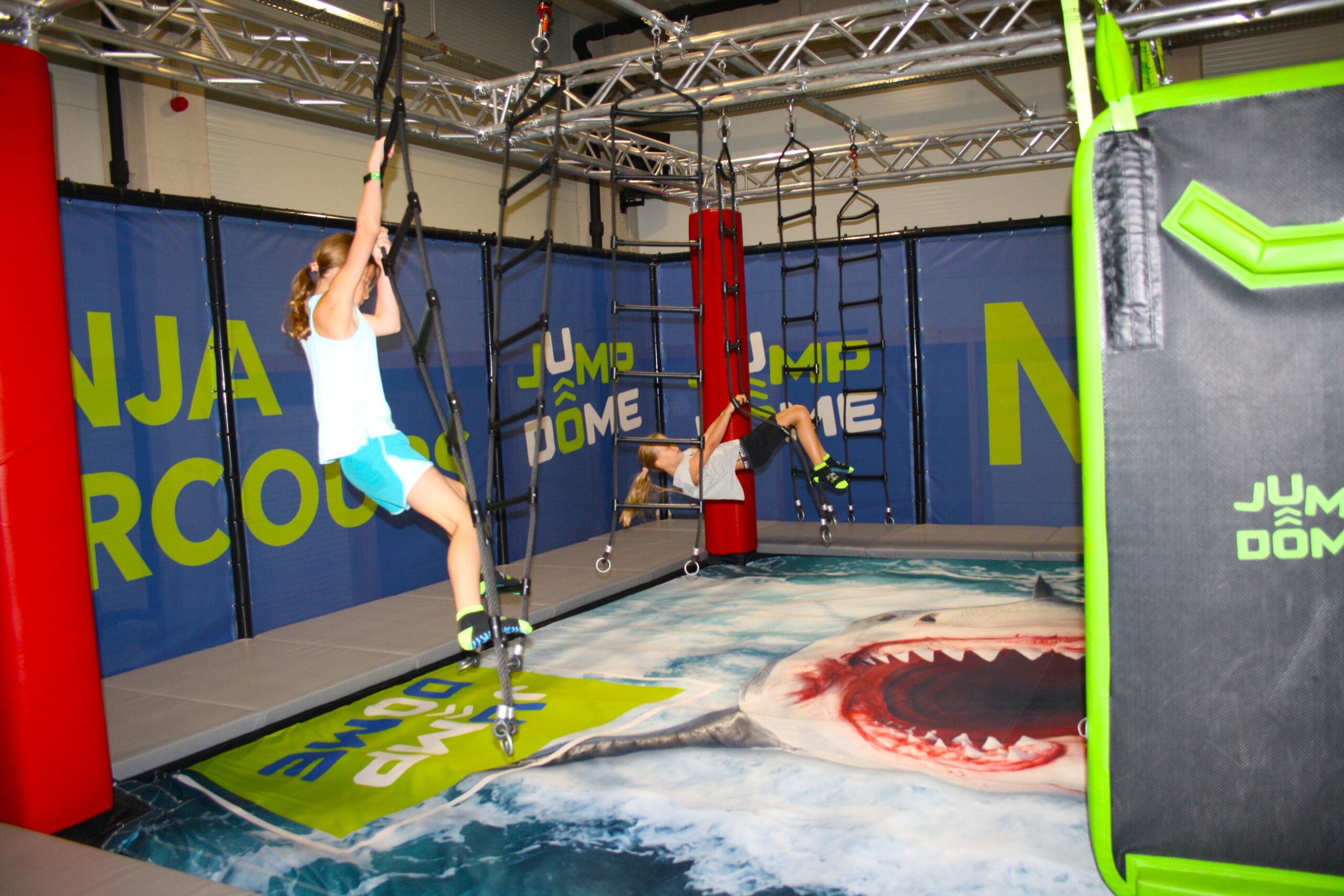 Zwei Personen klettern in einer Ninja-Parkour-Halle an h&auml;ngenden Seilen &uuml;ber eine Matte mit Hai-Motiv.