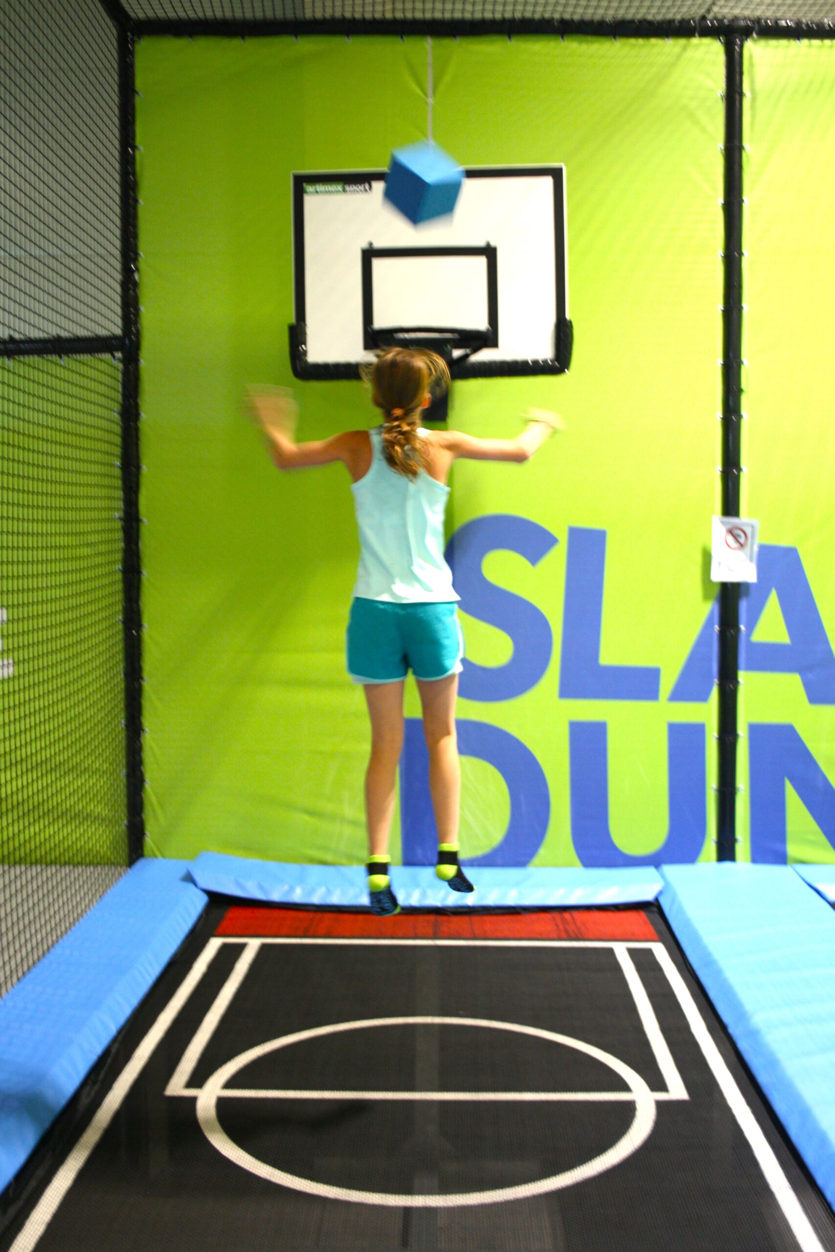 Ein M&auml;dchen springt auf einem Trampolin und zielt mit einem blauen Block auf einen Basketballkorb an einer gr&uuml;nen Wand.