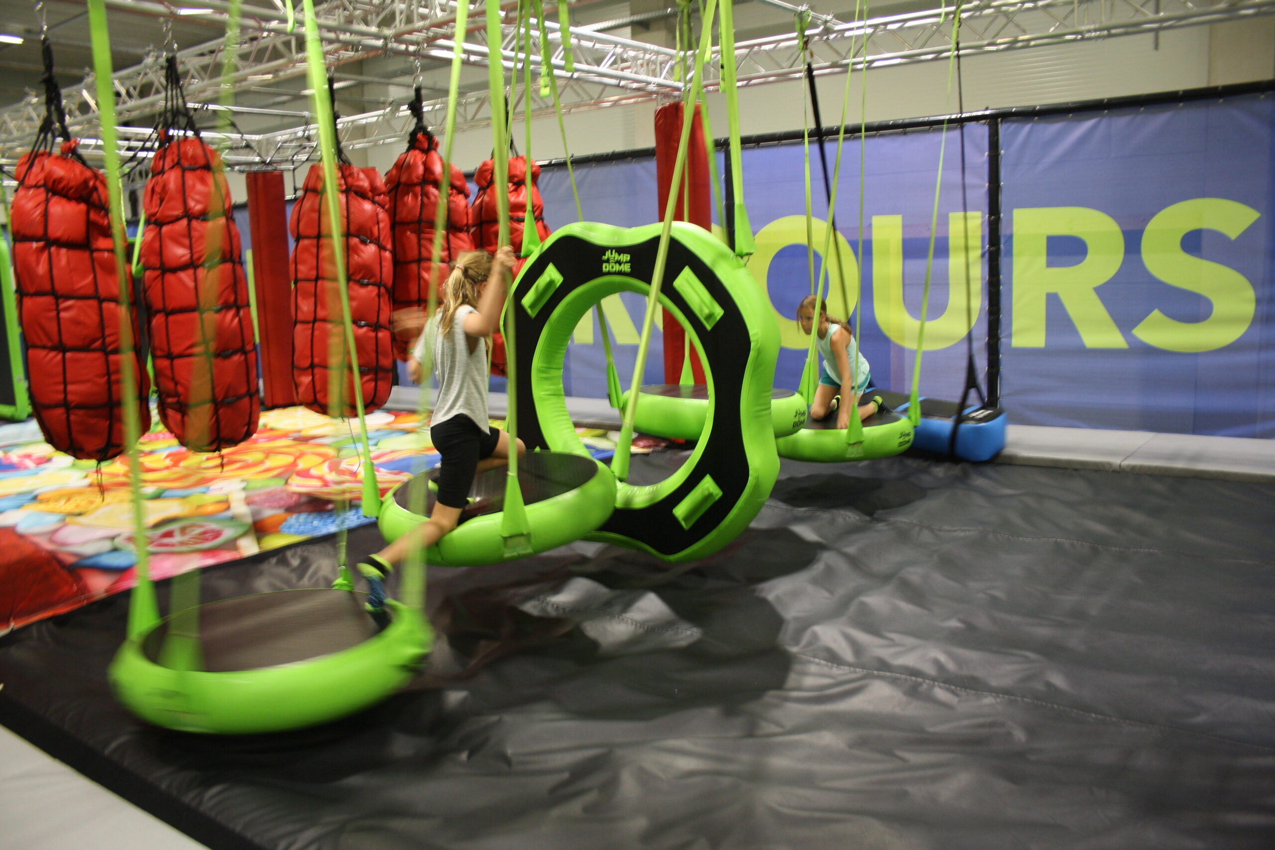 Zwei Kinder spielen auf gr&uuml;nen Schaukelringen in einem Indoor-Hindernisparcours mit gepolstertem Boden und gepolsterten W&auml;nden.