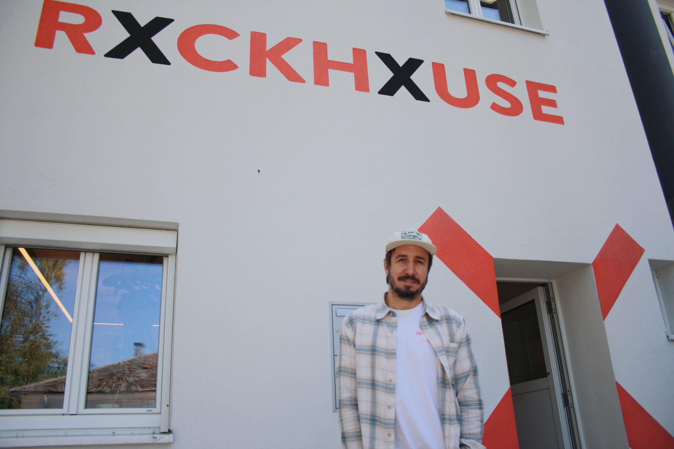Ein Mann in einem karierten Hemd und mit einem Hut steht vor einem Gebäude mit der Aufschrift „RXCKHXUSE“.