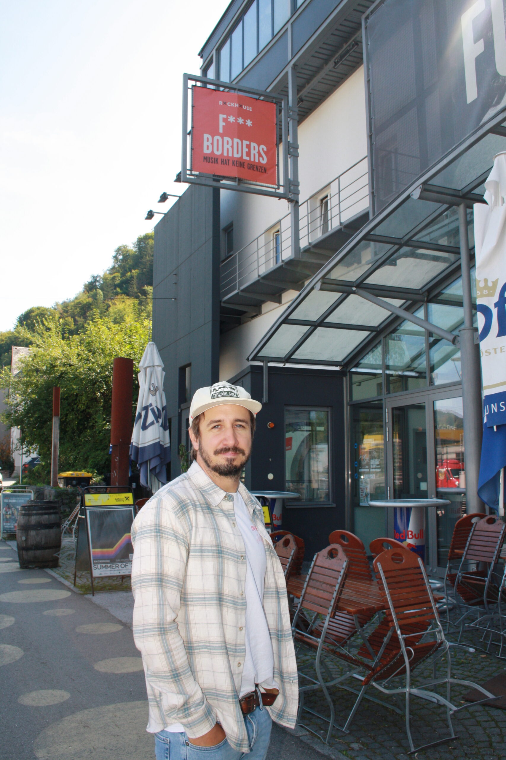 Ein Mann in einem karierten Hemd und einer M&uuml;tze steht vor einem Geb&auml;ude, &uuml;ber dem ein Schild mit der Aufschrift &bdquo;F*** BORDERS&ldquo; h&auml;ngt.