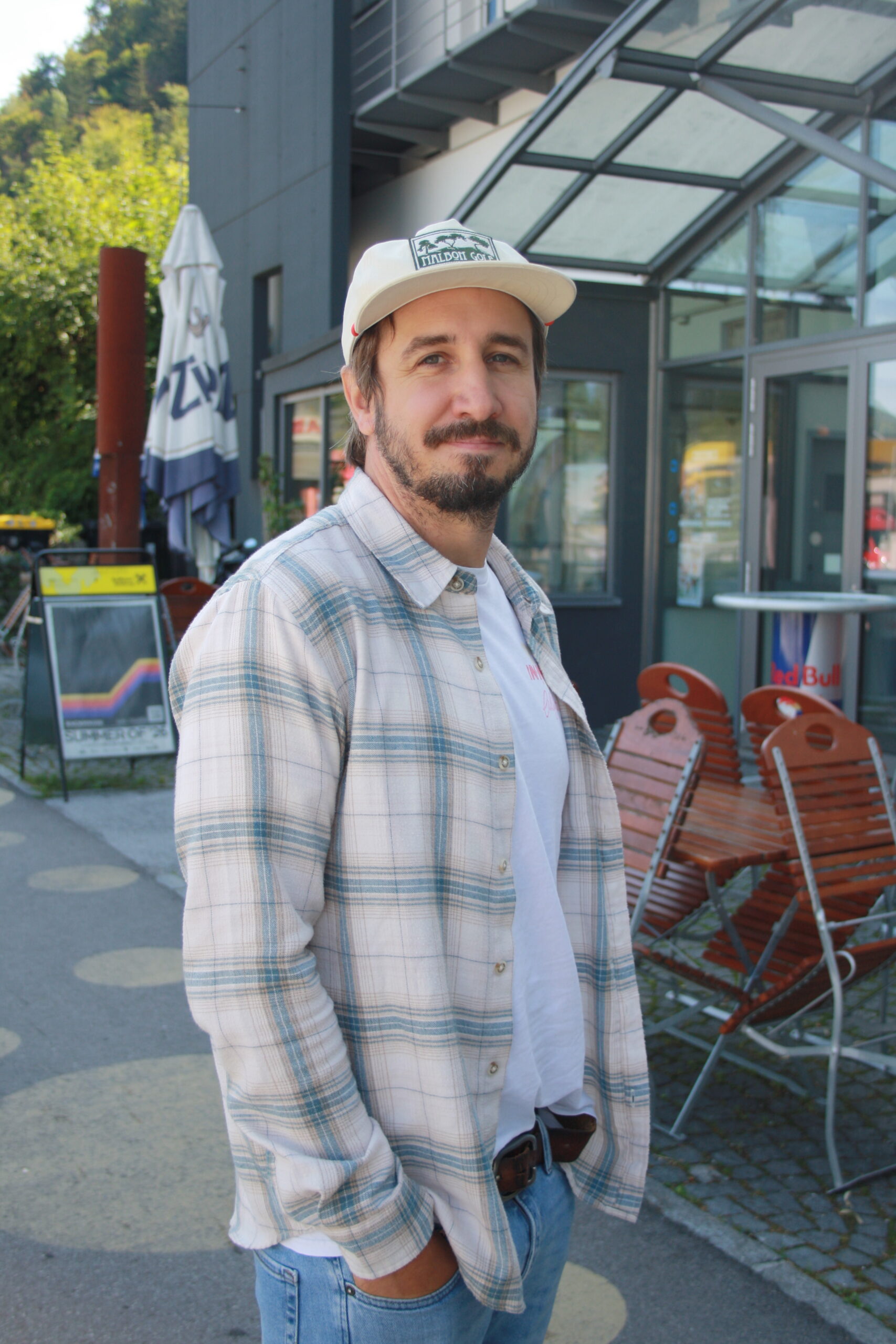 Ein Mann in einem karierten Hemd und einer M&uuml;tze steht vor einem Caf&eacute;, hinter ihm stehen St&uuml;hle und ein verglastes Eingangstor.