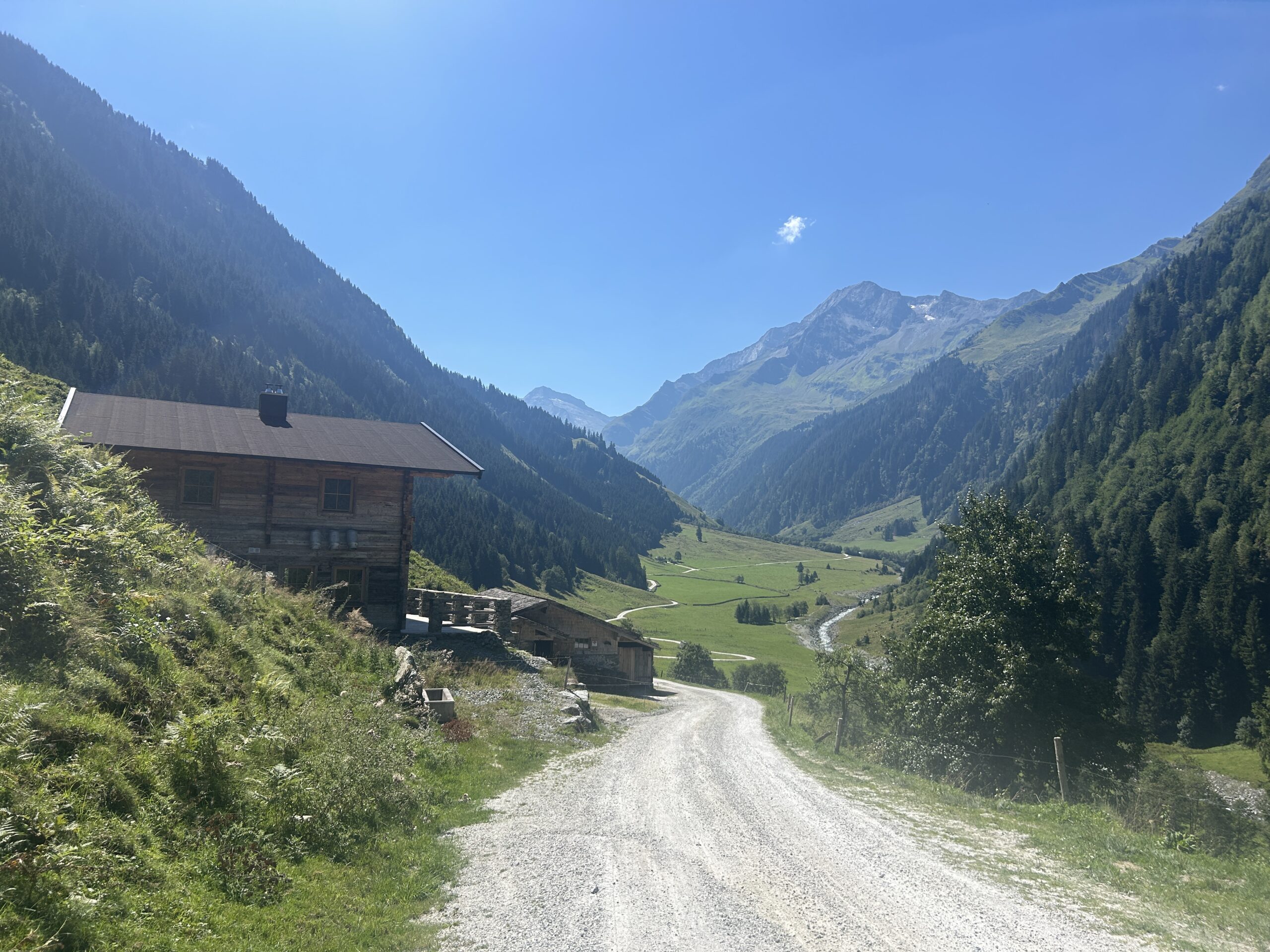 Eine Schotterstraße führt an einer Hütte vorbei durch ein grünes Tal, umgeben von Bergen unter einem strahlend blauen Himmel.