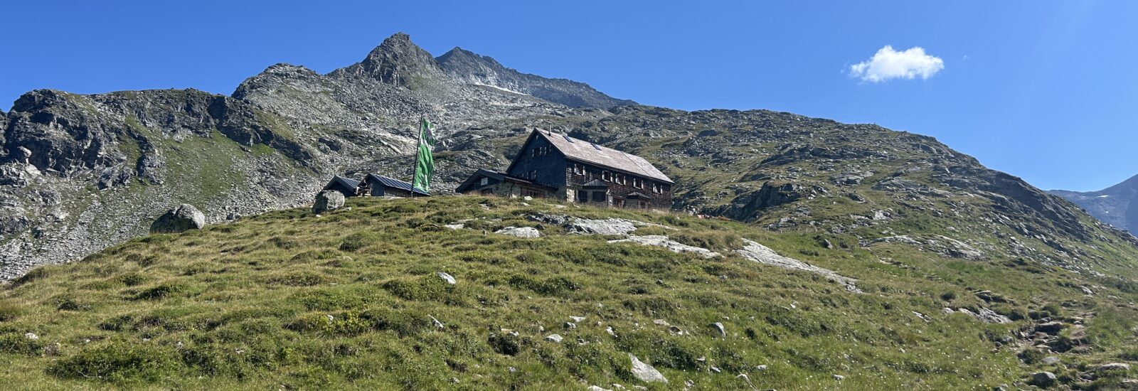 Eine Berghütte mit einer grünen Fahne steht an einem grasbewachsenen Hang unter einem strahlend blauen Himmel und felsigen Gipfeln.